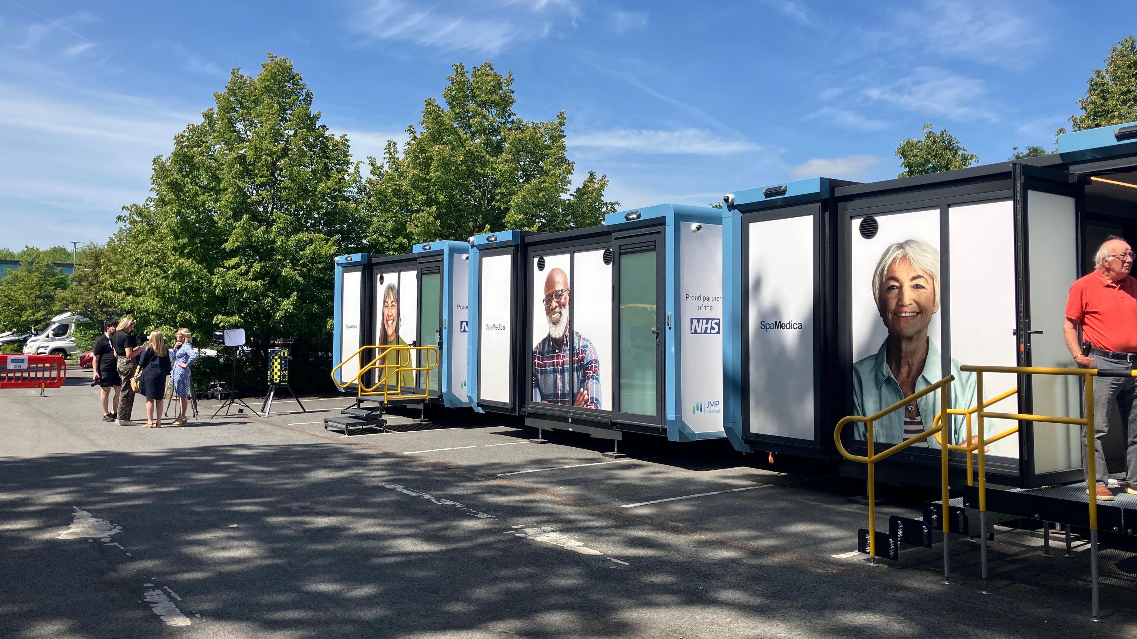 Three white demountable buildings in a sunny car park with a handful of people with grey hair stood outside.