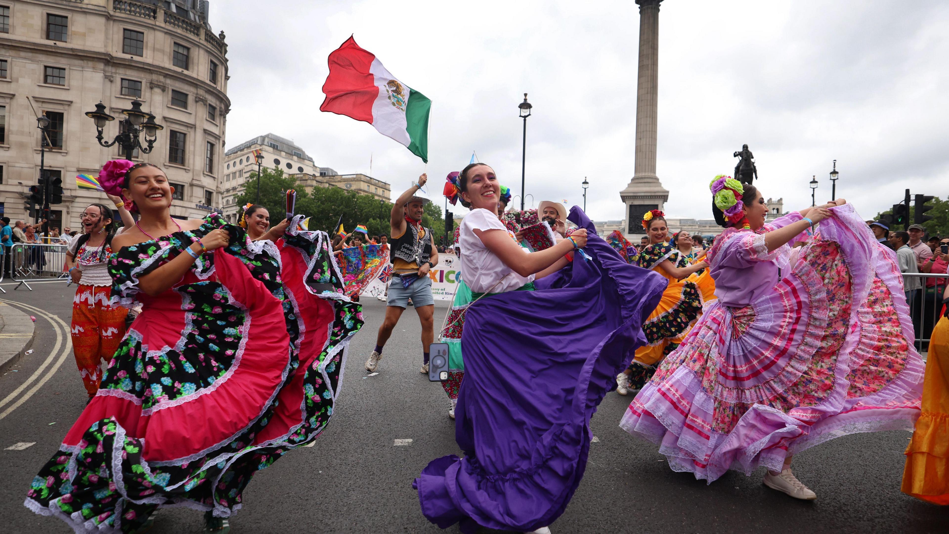 Three people in traditional Mexican dresses lift up their skirts as they dance through Trafalgar Square in central London. 
