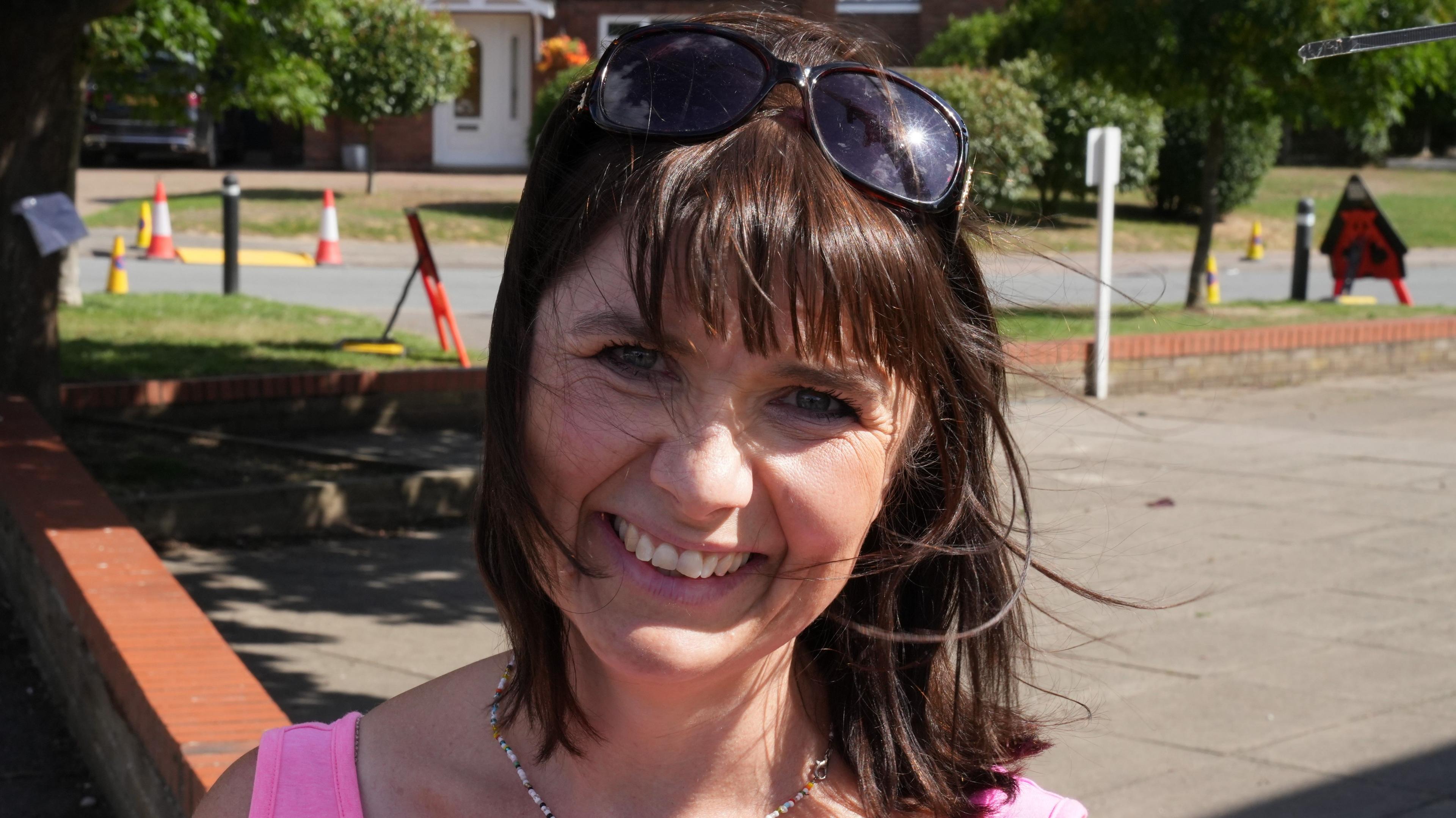 A woman smiles at the camera. She has short brown hair with sunglasses resting on top of her head. She wears a bright pink vest top.