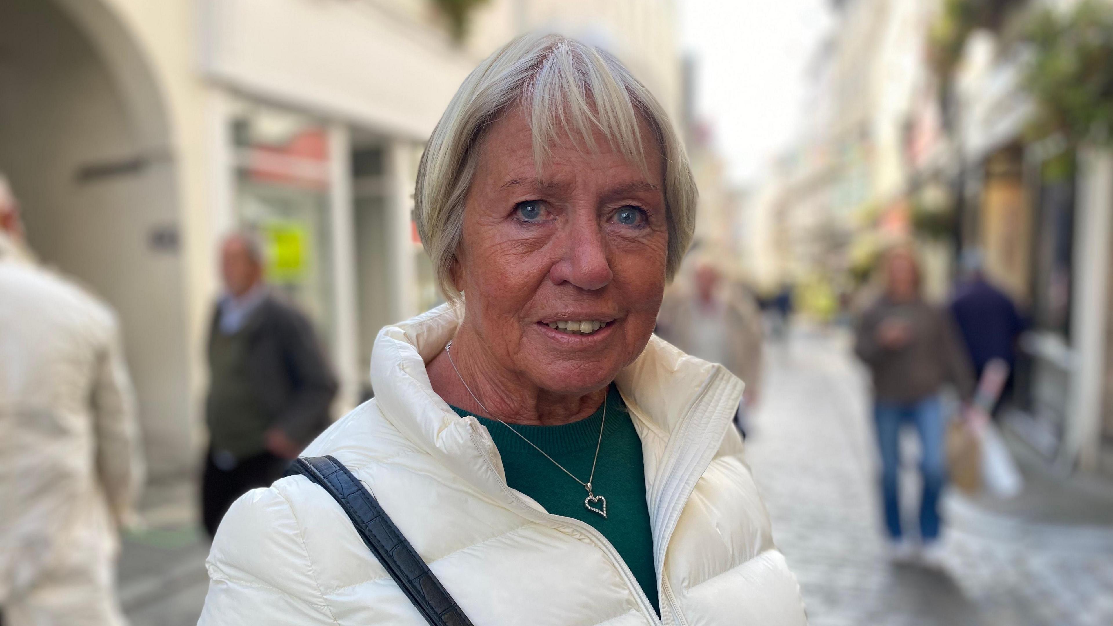 An older woman with short, blonde hair in a cream puffer jacket is pictured standing in a cobbled high street, with a smattering of shoppers in the background. 