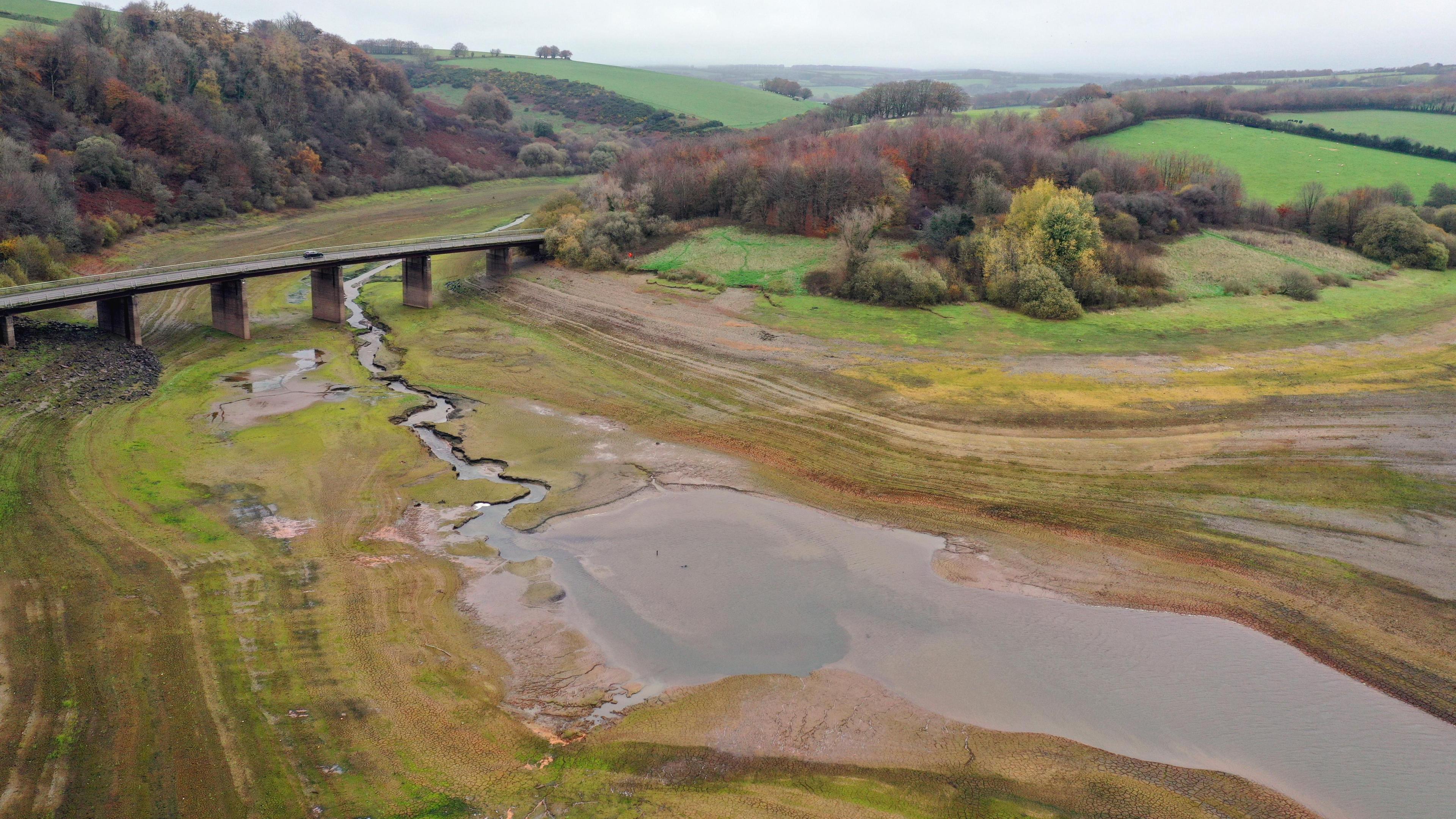 An aerial view of Wimbleball Reservoir on Exmoor with large patches of grass and mud visible and large pools of water. A bridge crossing the reservoir can be seen, along with areas of woodland around the edge