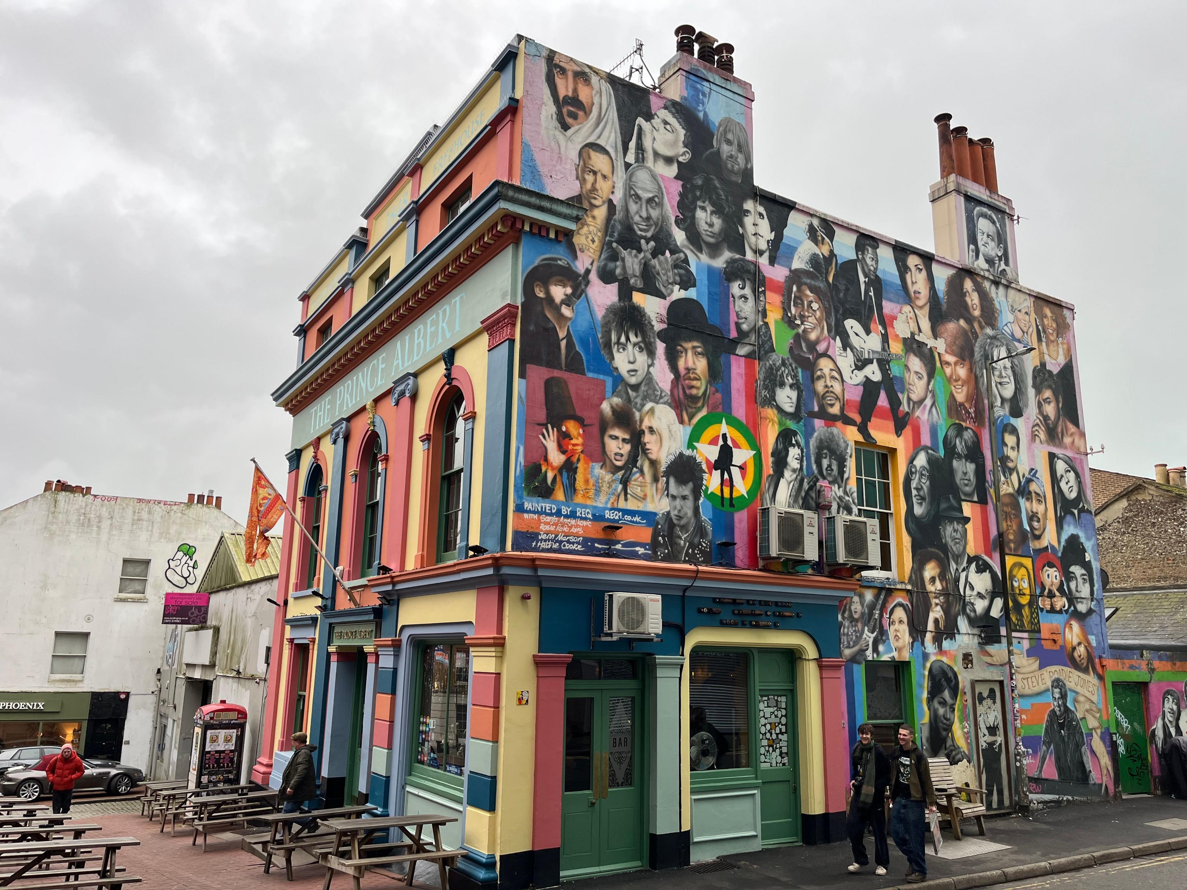 The Prince Albert building which is multicoloured in yellow, blue, green, orange and red paint. One wall is covered with a mural of celebrities including Elvis Presley, Eminem and Amy Winehouse. There is also a Banksy piece of art called Kissing Coppers. There is a large Prince Albert logo on the front and wooden benches outside the front and a flag hanging down