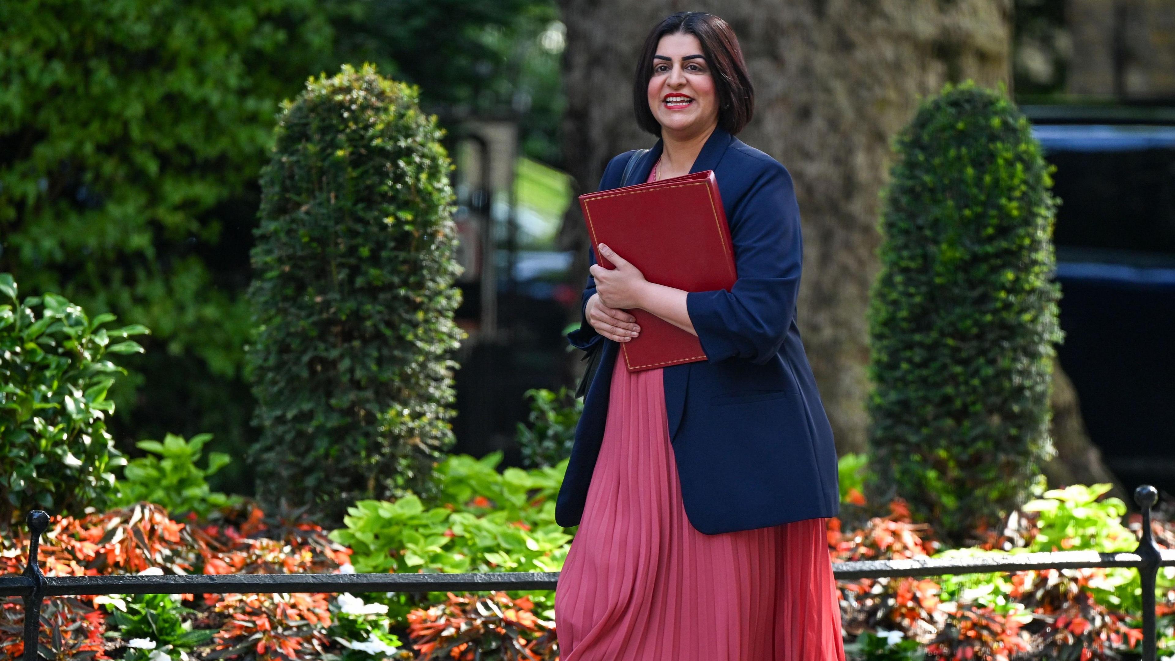 Shabana Mahmood, UK home secretary, arrives for a cabinet meeting at 10 Downing Street in London in a pink dress with a navy blazer, while holding a red box and smiling. Green foliage is seen behind her.