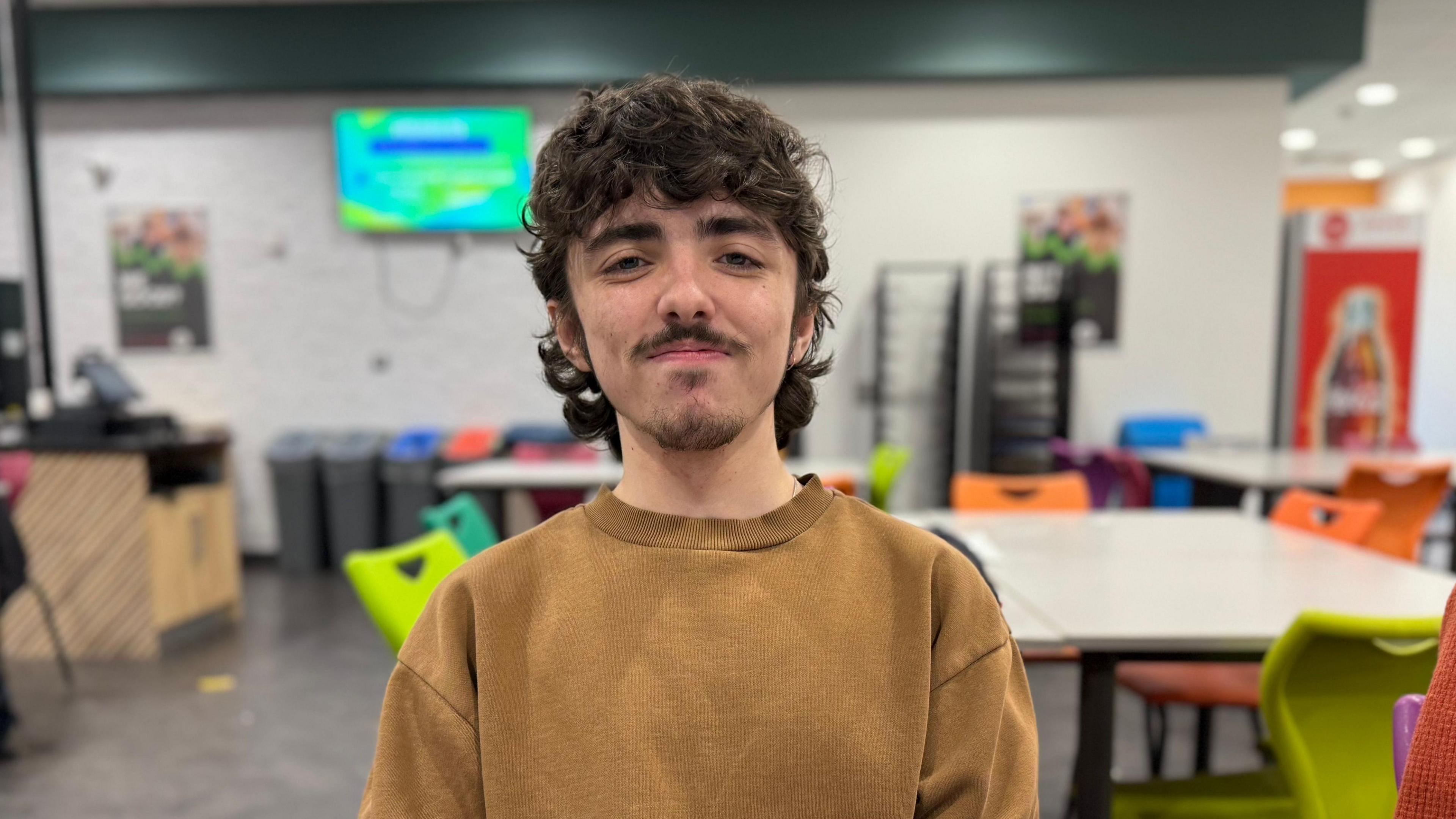 Cavan Griffin sits at a table within a food canteen at Coleg y Cymoedd in Nantgarw. He has a brown curly mullet and a goatie beard and moustache. He smiles at the camera, a television and tables are behind him. 