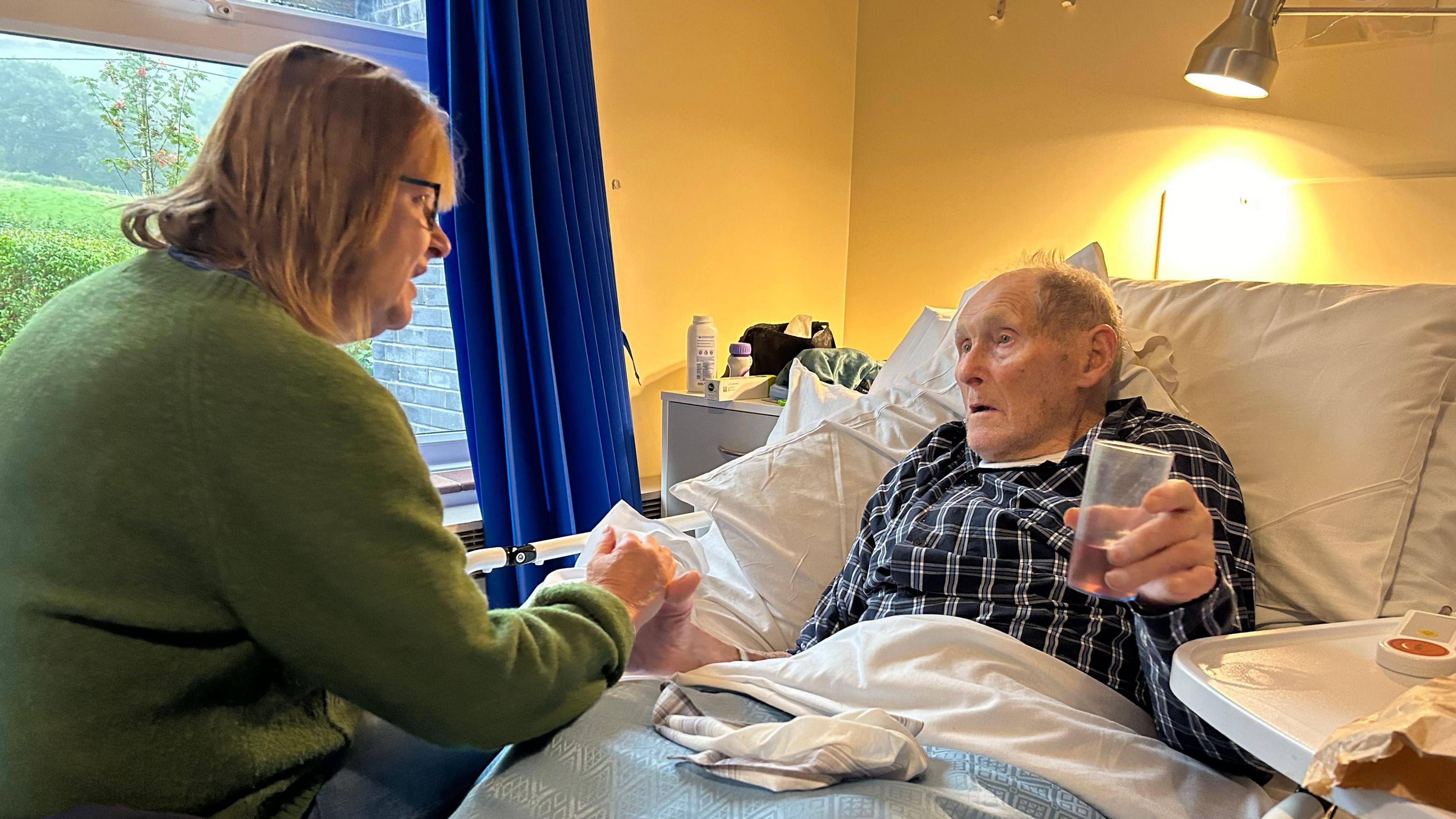 Rhythwyn Evans in a hospital bed, wearing black check pyjamas and holding the hand of a woman in a green cardigan while he holds a plastic cup of squash in the other.