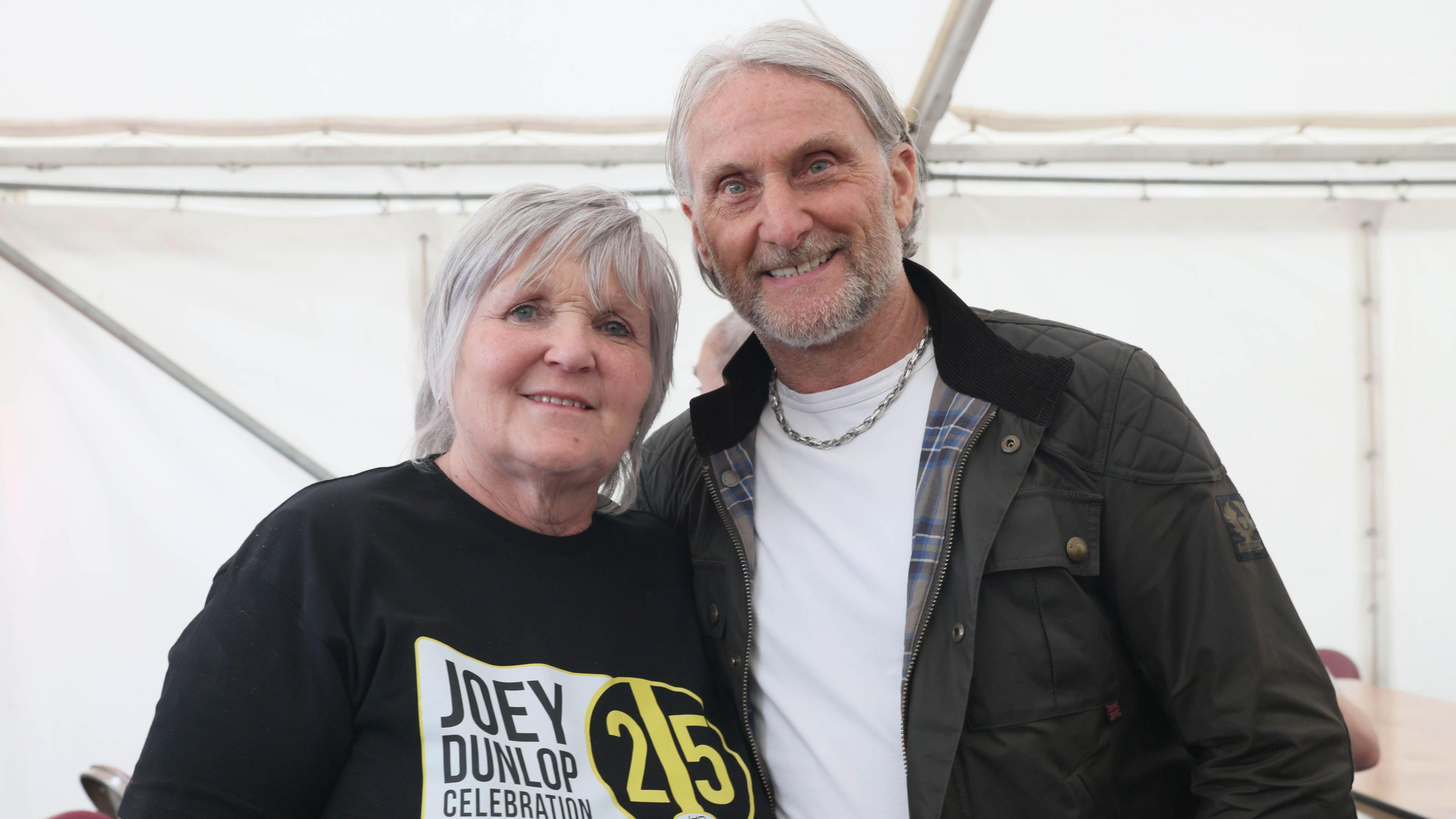 Linda, left, has short grey hair and is smiling at the camera. She is wearing a black t-shirt which says Joey Dunlop Celebration with the number 25 in yellow. She is standing next to Carl Fogarty. They are hugging into each other. Carl has longish grey hair pushed behind his ears. He is wearing a green coat with blue tartan lining and a white t-shirt. He is smiling at the camera.