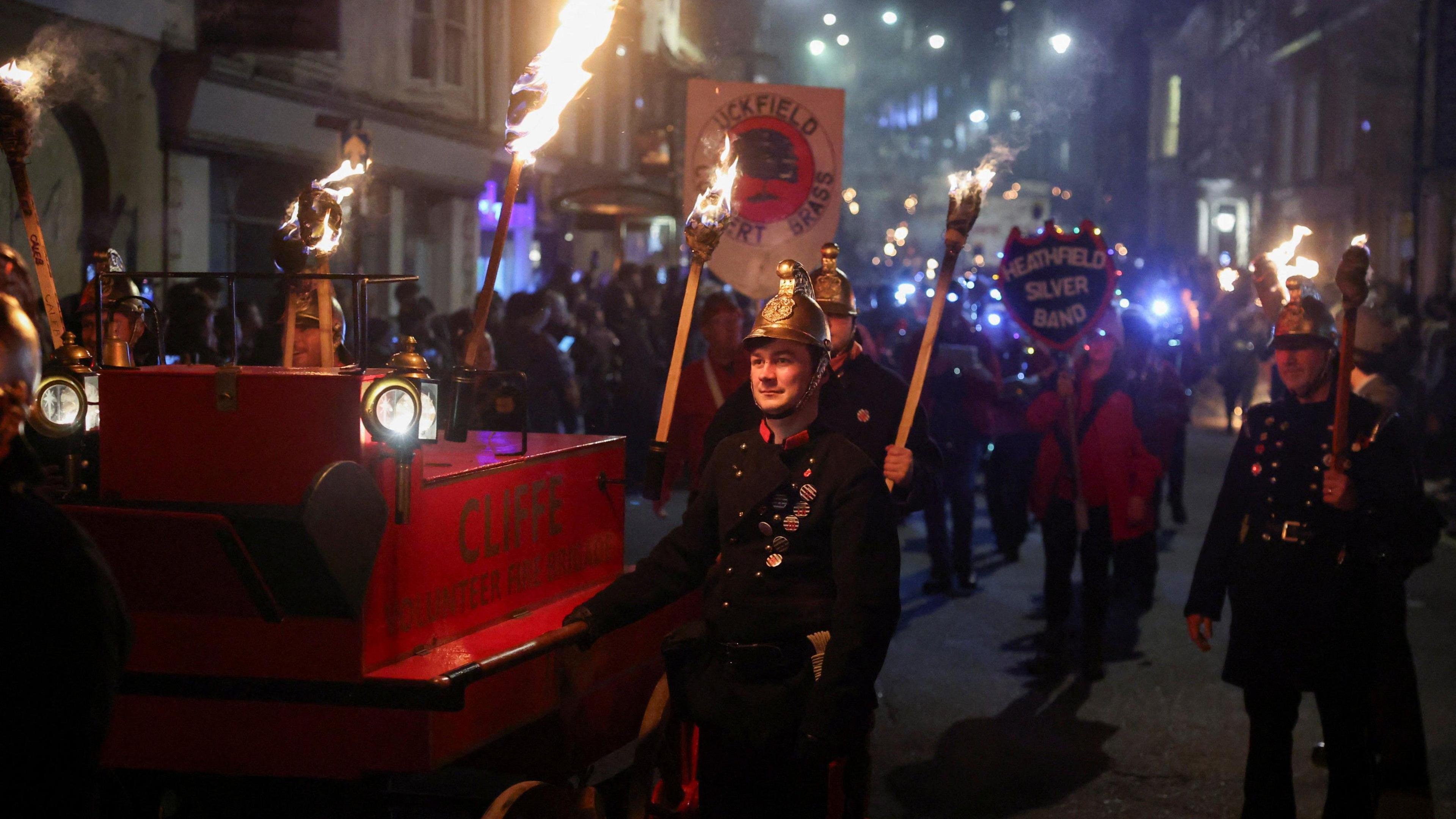People wearing Victorian fire fighter outfits and carrying burning torches walk alongside a vintage fire engine.
