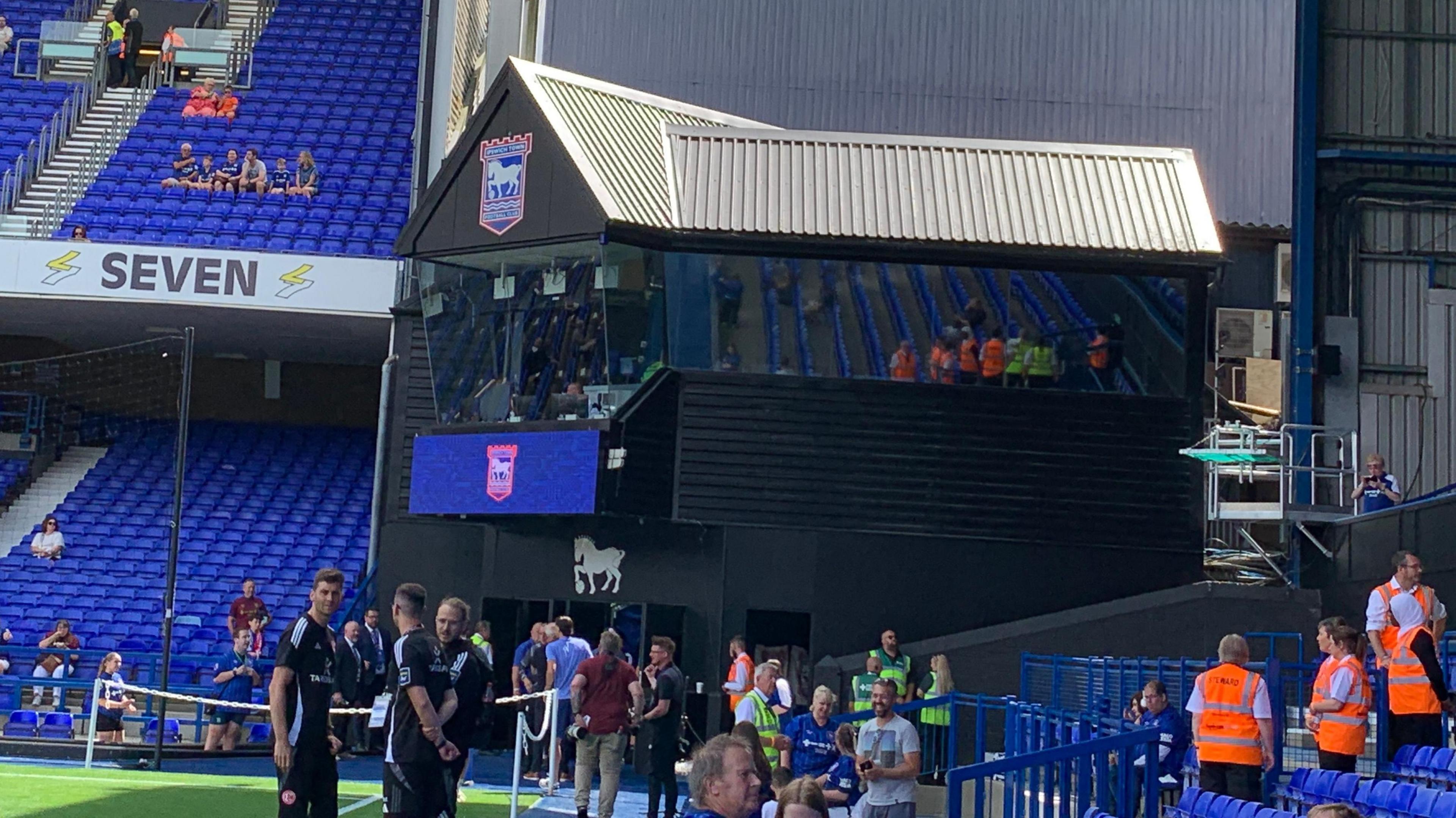 A black, two-tier building with a glass facade in a corner of Portman Road. It has the Ipswich Town FC crest on it and lots of people standing around it. There are stands either side of the building, all with blue seats.