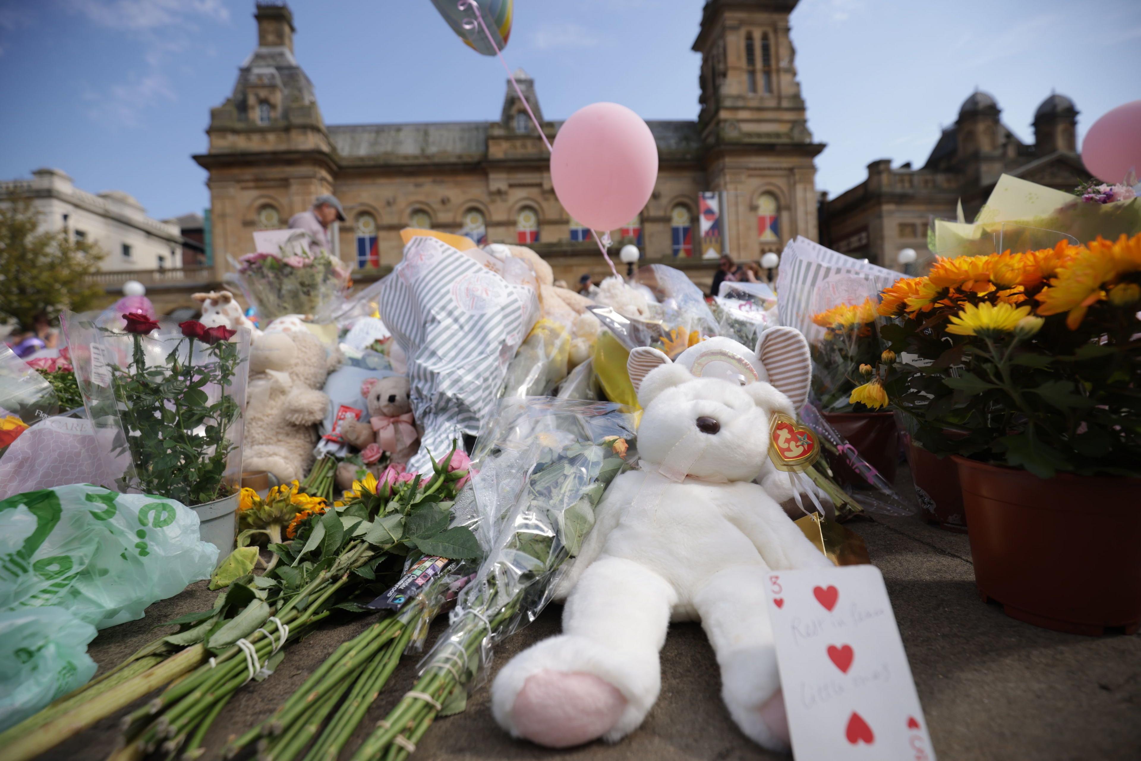 A teddy bear and flowers sit on the ground in a memorial