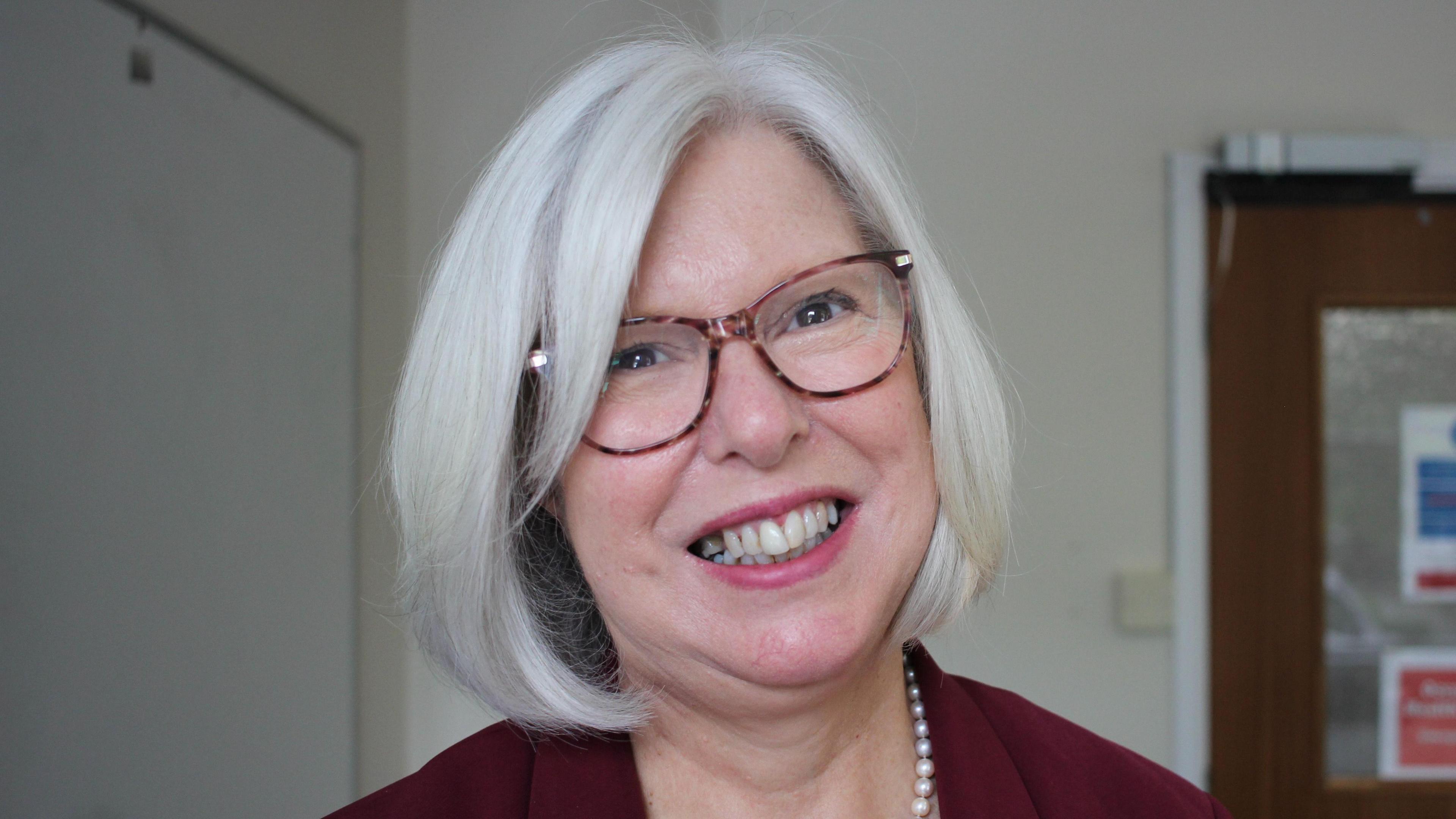 A woman with white hair and tortoiseshell glasses smiles at the camera, she is wearing pears and a maroon blazer. Behind her is a brown office door. 