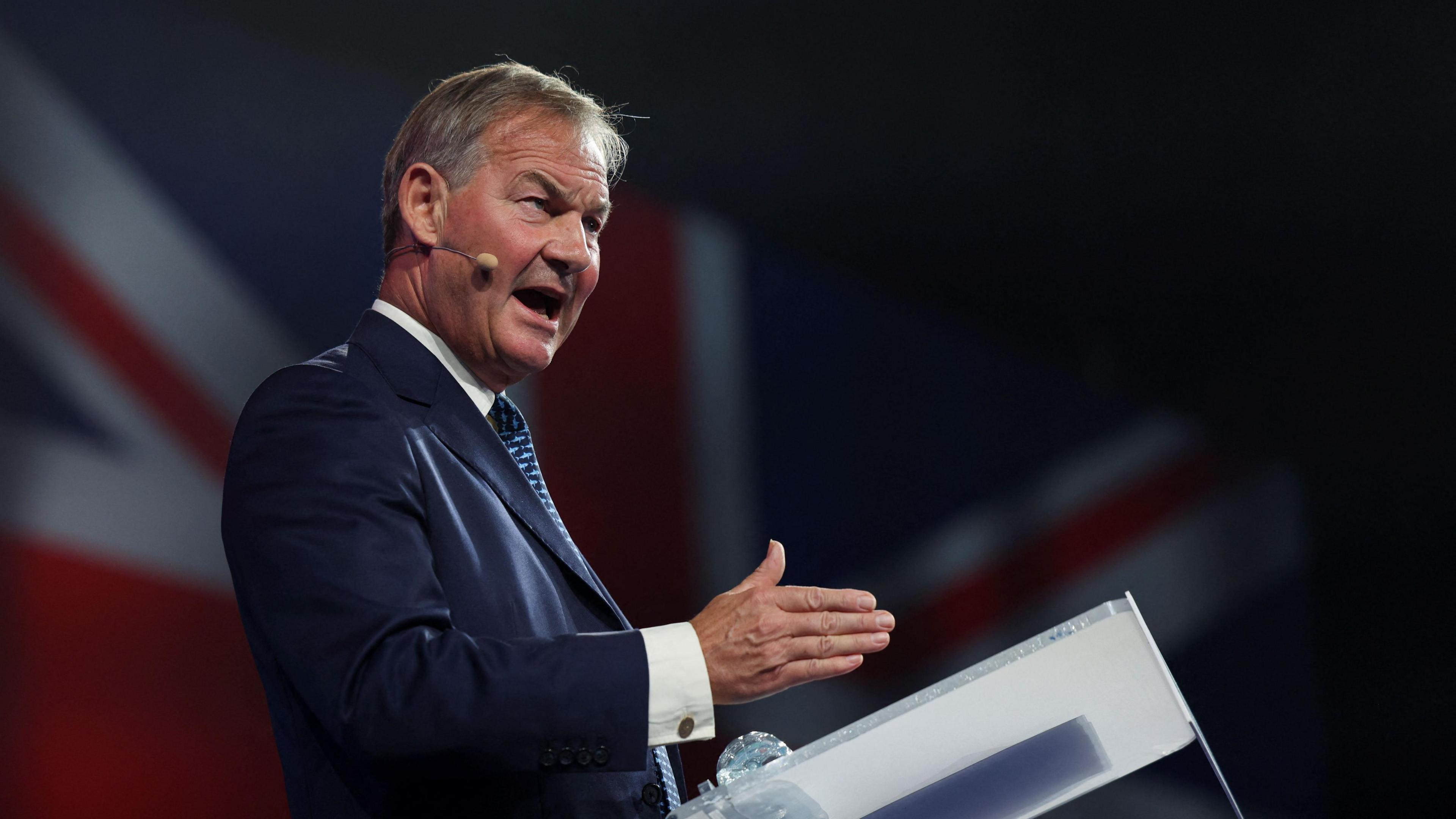 Rupert Lowe is standing at a podium giving a speech. He's wearing a dark blue jacket, white shirt and blue striped tie. Behind him, we can see a Union Flag.