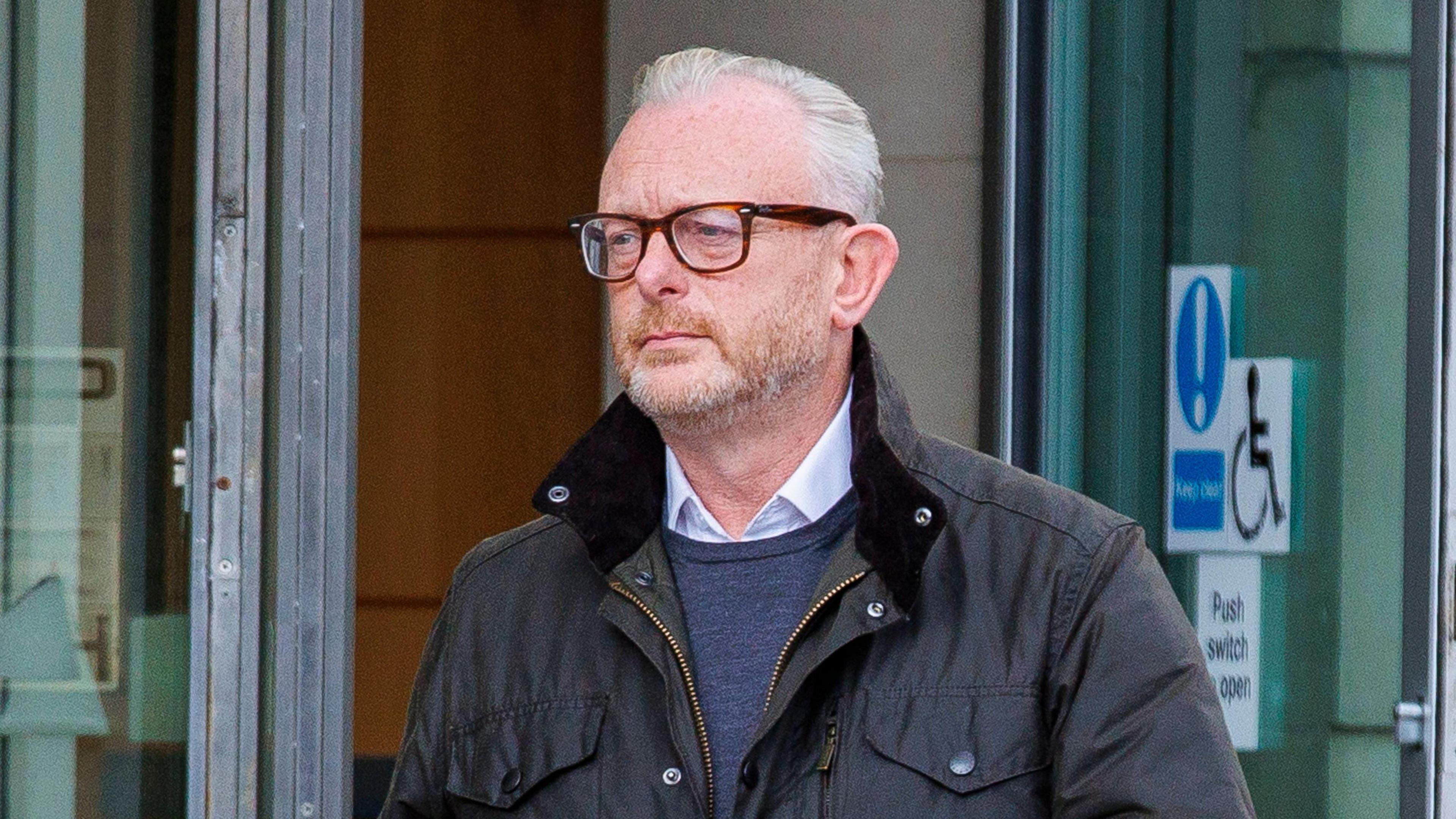 A man with short grey hair is pictured walking out of a building. He is wearing brown rimmed glasses, a white shirt, blue jumper and dark coloured coat.