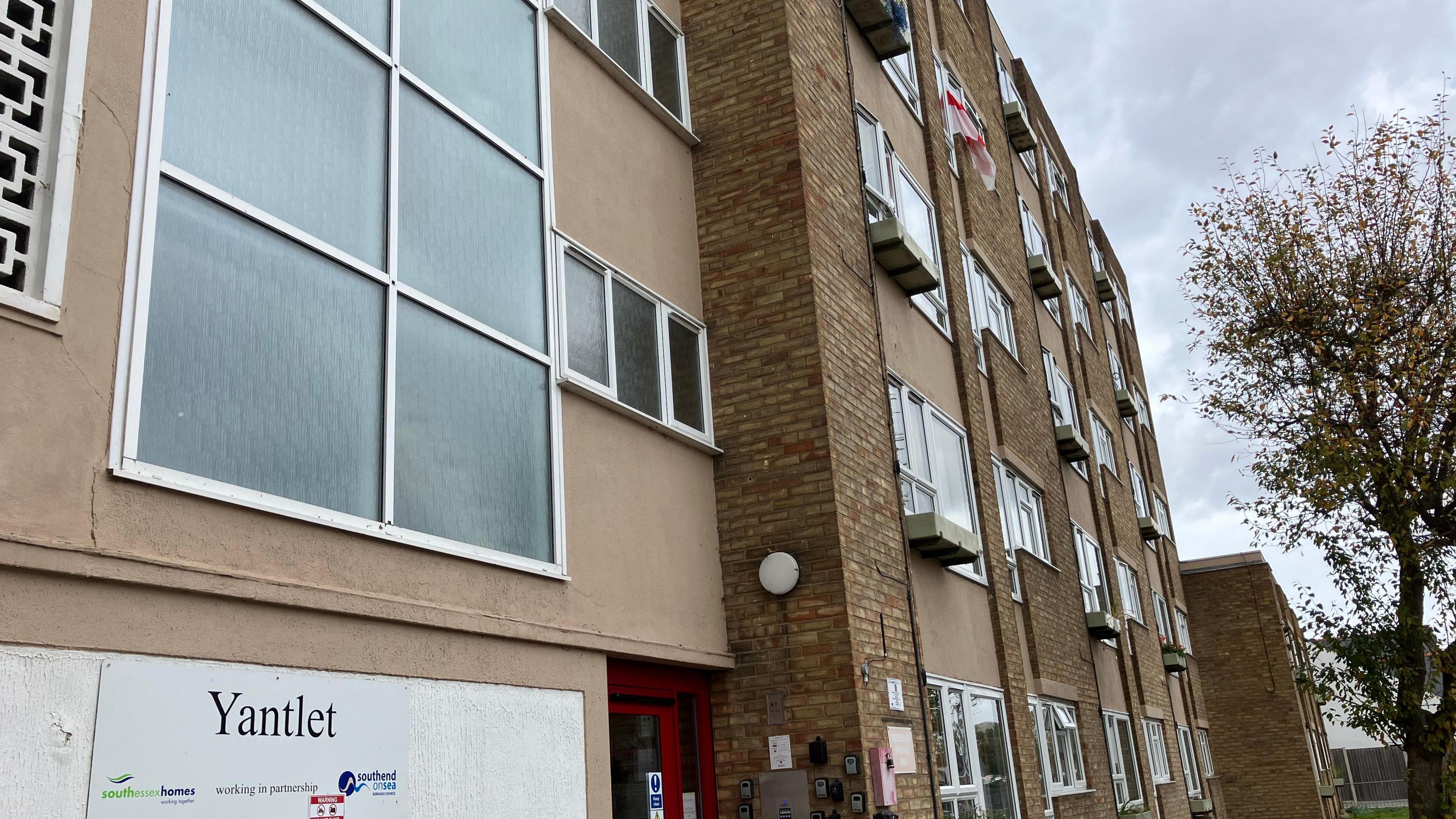 Yantlet House is a beige tower block, with balcony ledges outside some of the flats. It is cloudy and there is a tree in the right of the frame.