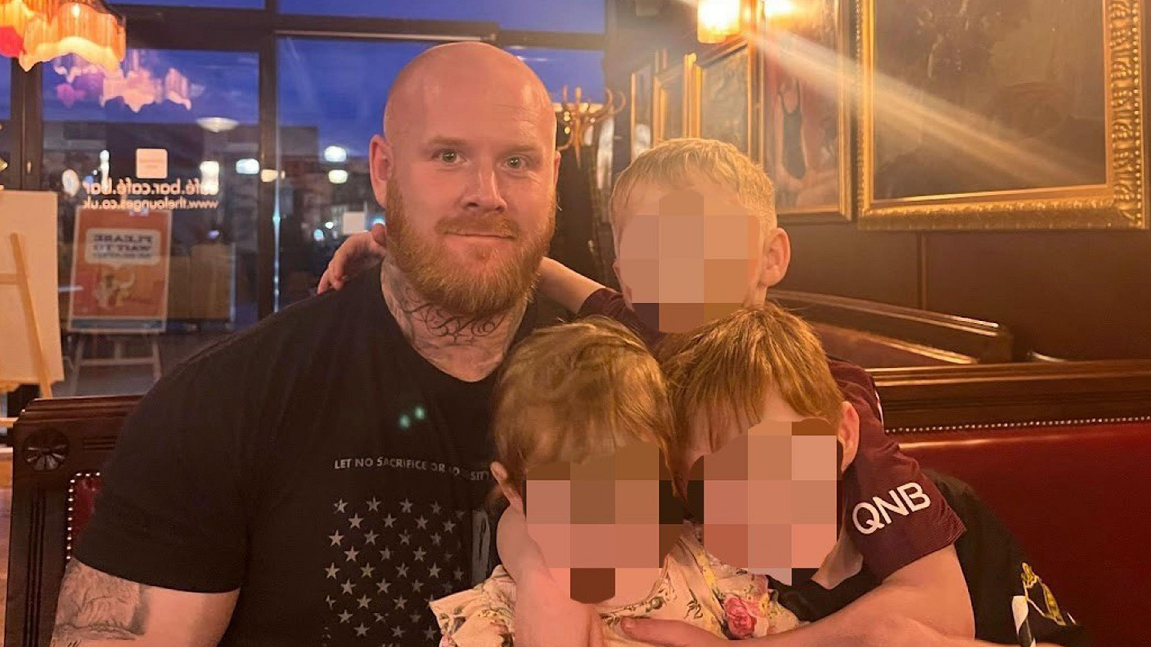 A family handout of Lenny Scott with all three of his children. He is wearing a black t-shirt and has his arms wrapped around the kids. They are sitting in a restaurant.