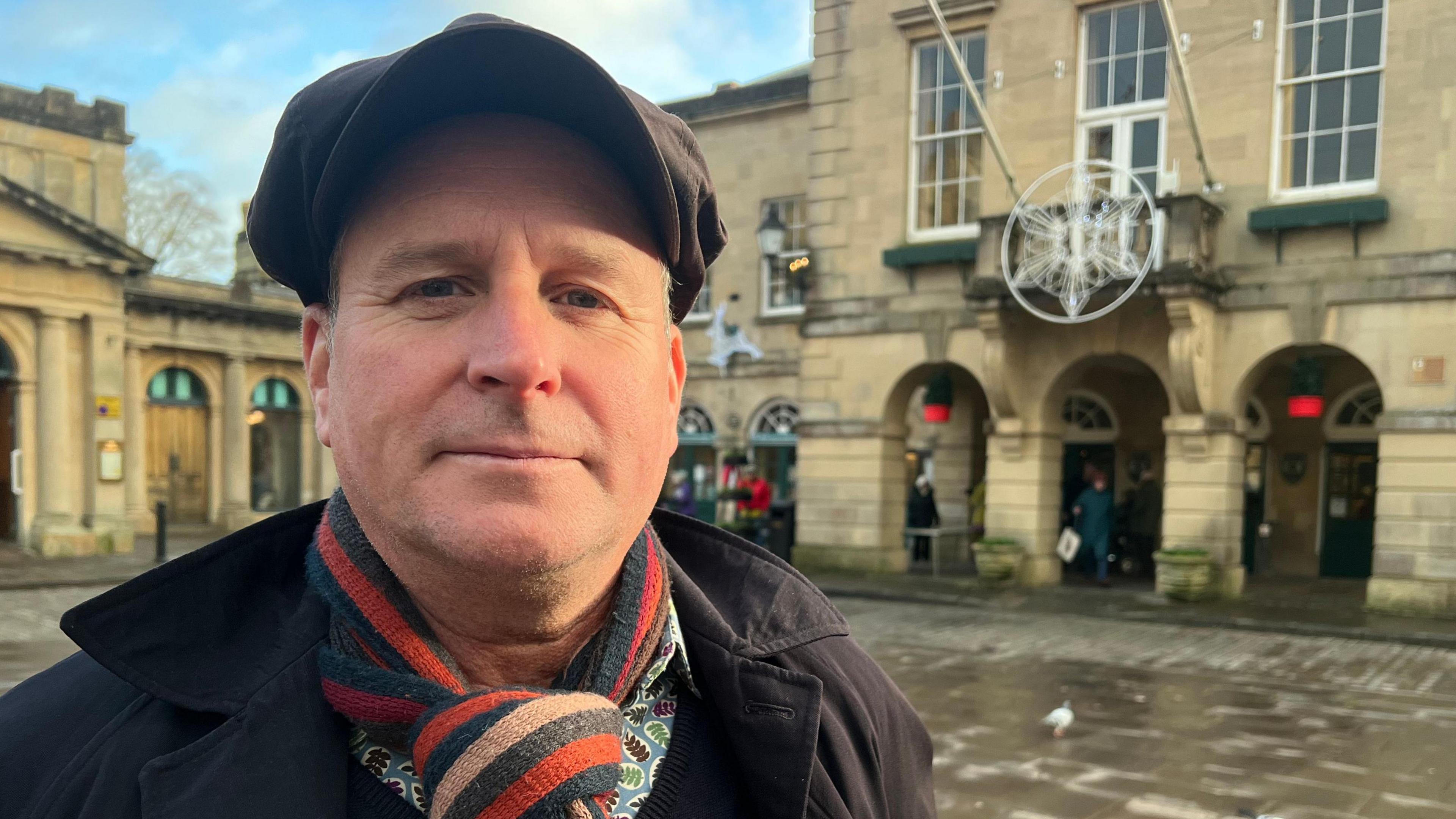 Mike looks into the camera wearing a black cap, black jacket and red, black and cream striped scarf.