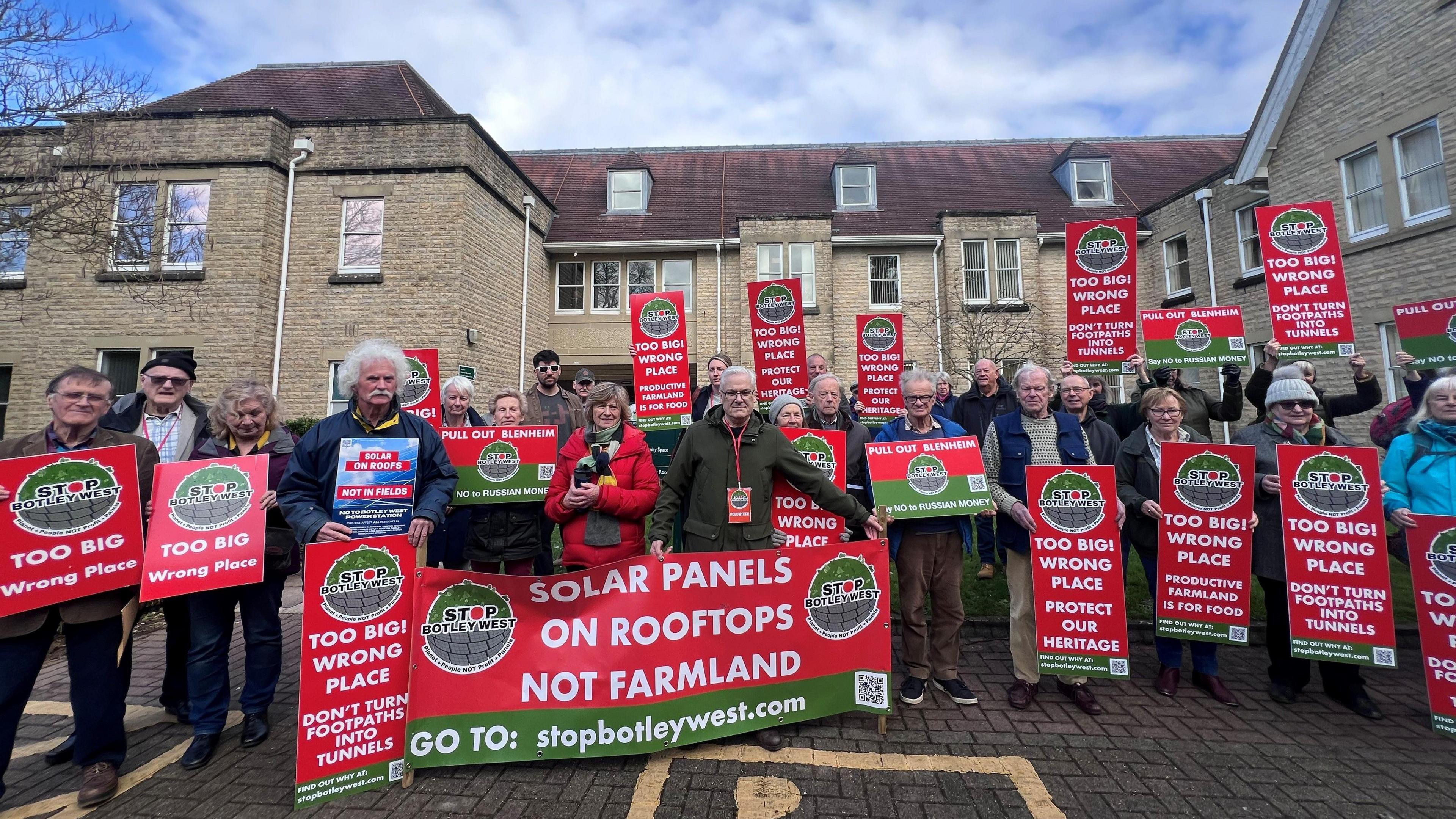 Protesters from the Stop Botley West group outside West Oxfordshire District Council's headquarters holding signs, most of which say: "Too big! Wrong place. Protect our heritage." 