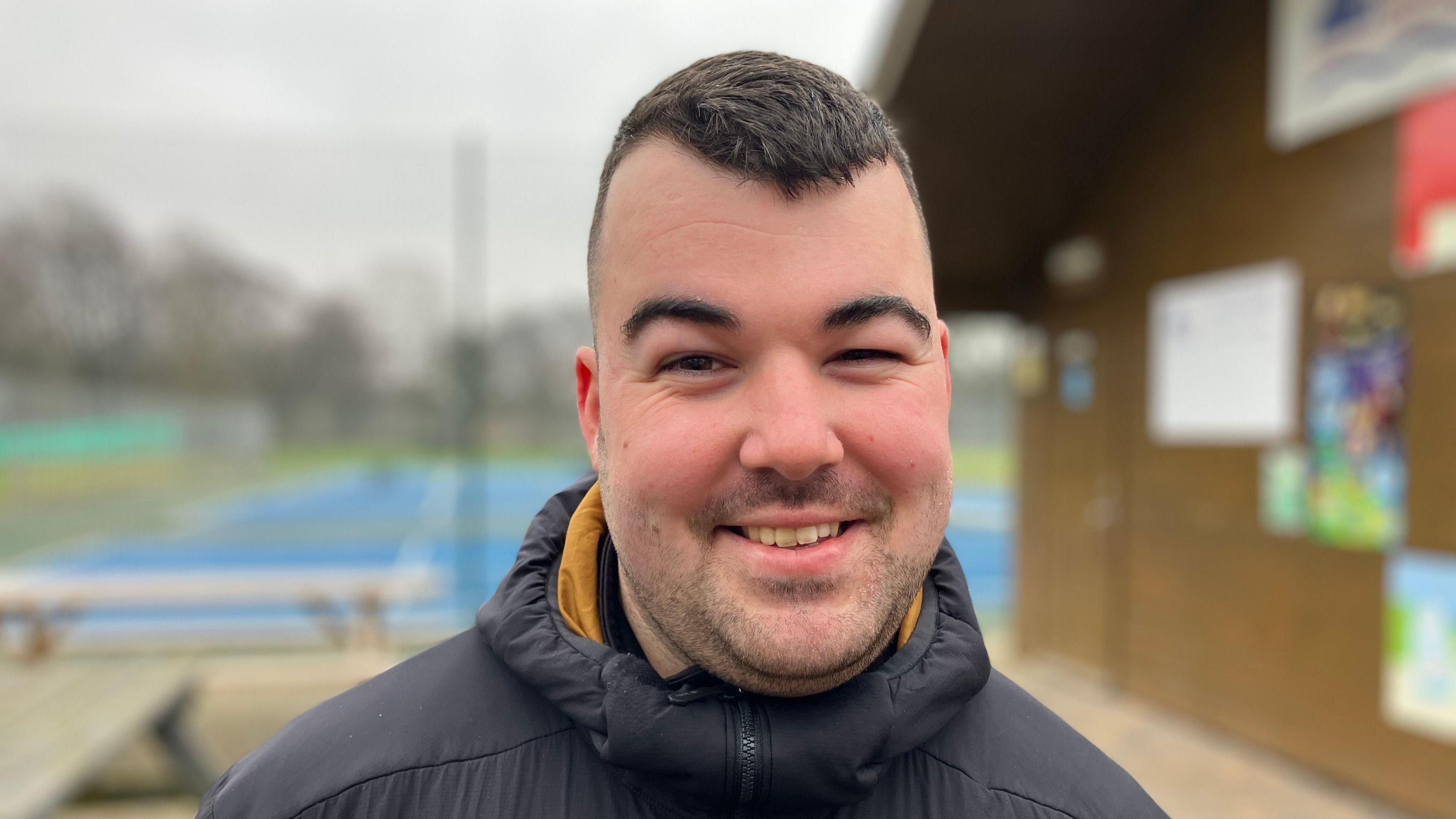 Ethan Edwards is at a sports centre wearing his coaching gear and smiling.