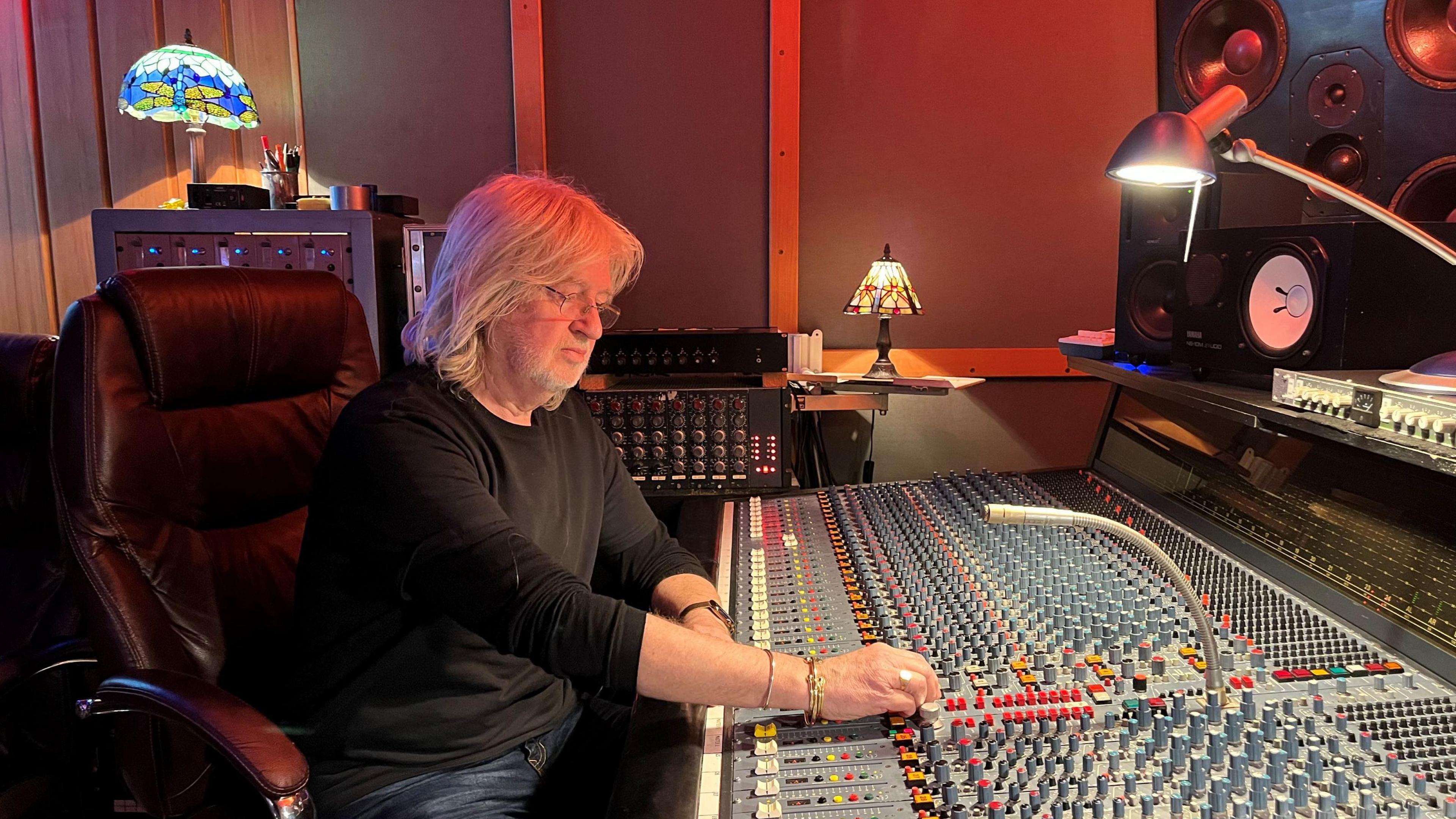 A man with shoulder-length blonde hair and glasses sitting in a large black leather chair. He is wearing a black long-sleeved top and jeans. He is leaning over a large sound mixing board and adjusting some of the dials.