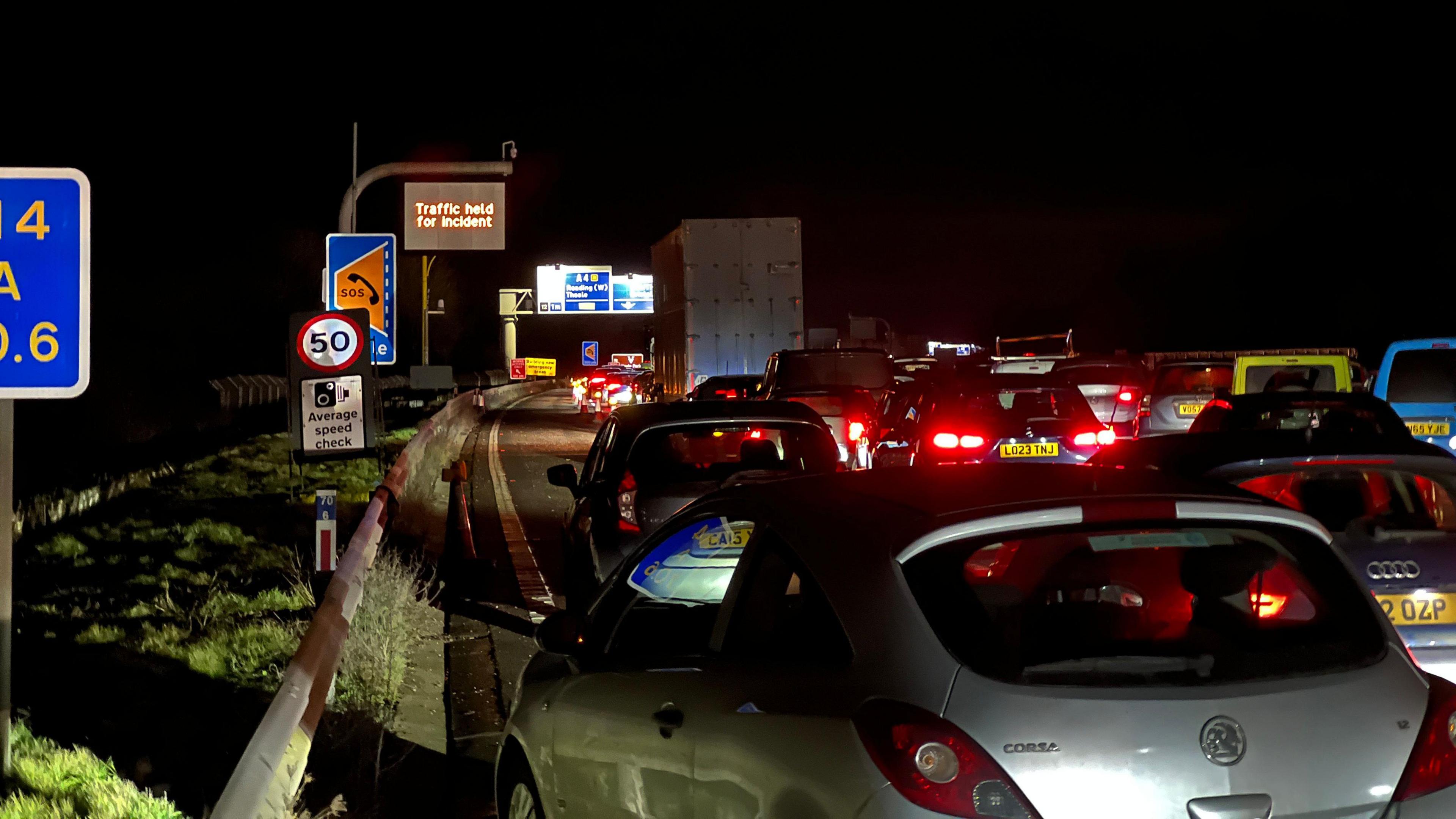 All three lanes of the M4 are full of cars at a standstill with their red breaklights on, there is a road sign to the left which says 'Traffic held for incident'