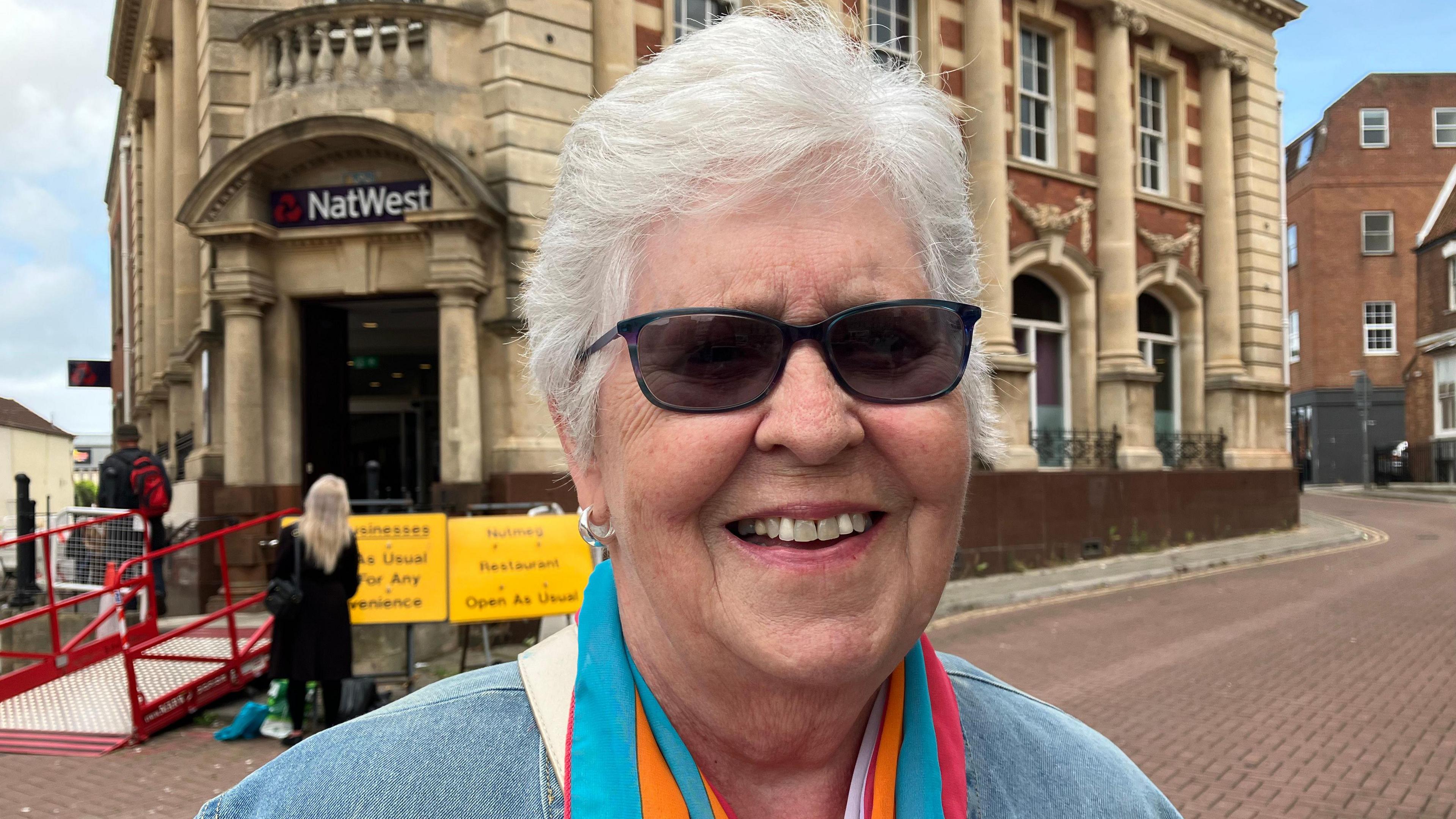 Headshot of a woman with white hair, smiling, wearing dark sunglasses and a colourful scarf standing outside a high street bank