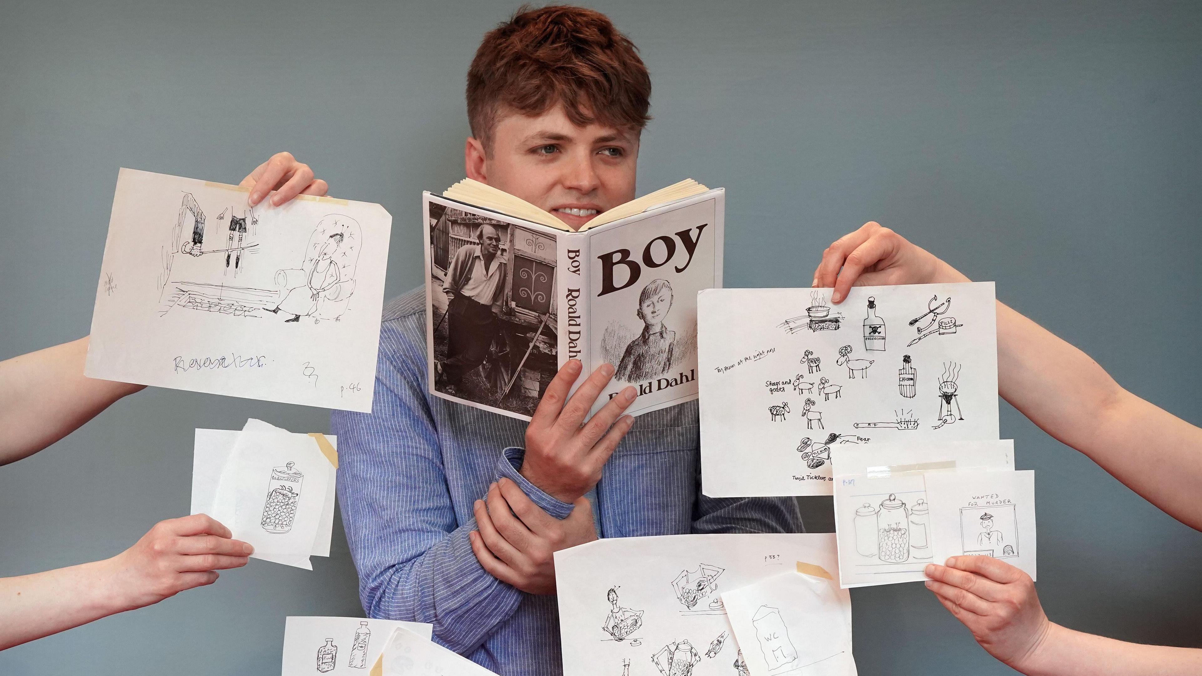 A man with brown hair smiles as he holds a copy of Boy with several people holding the illustrations from the book around him.