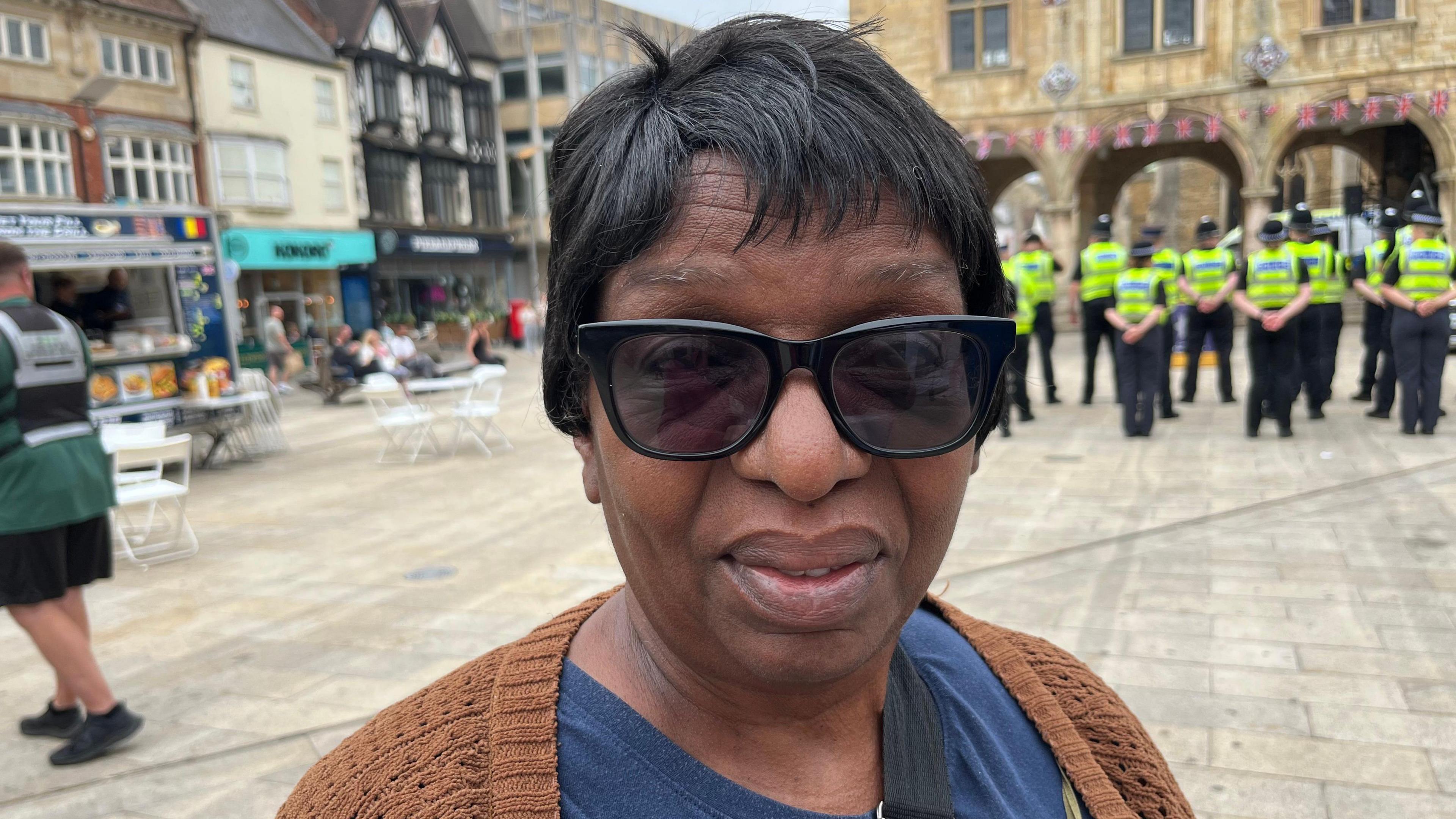 Woman wearing dark glasses stands in Peterborough square. She has black hair and looks at the camera. Behind her are lots of police officers and members of the public. A church can be seen in the background and shops.