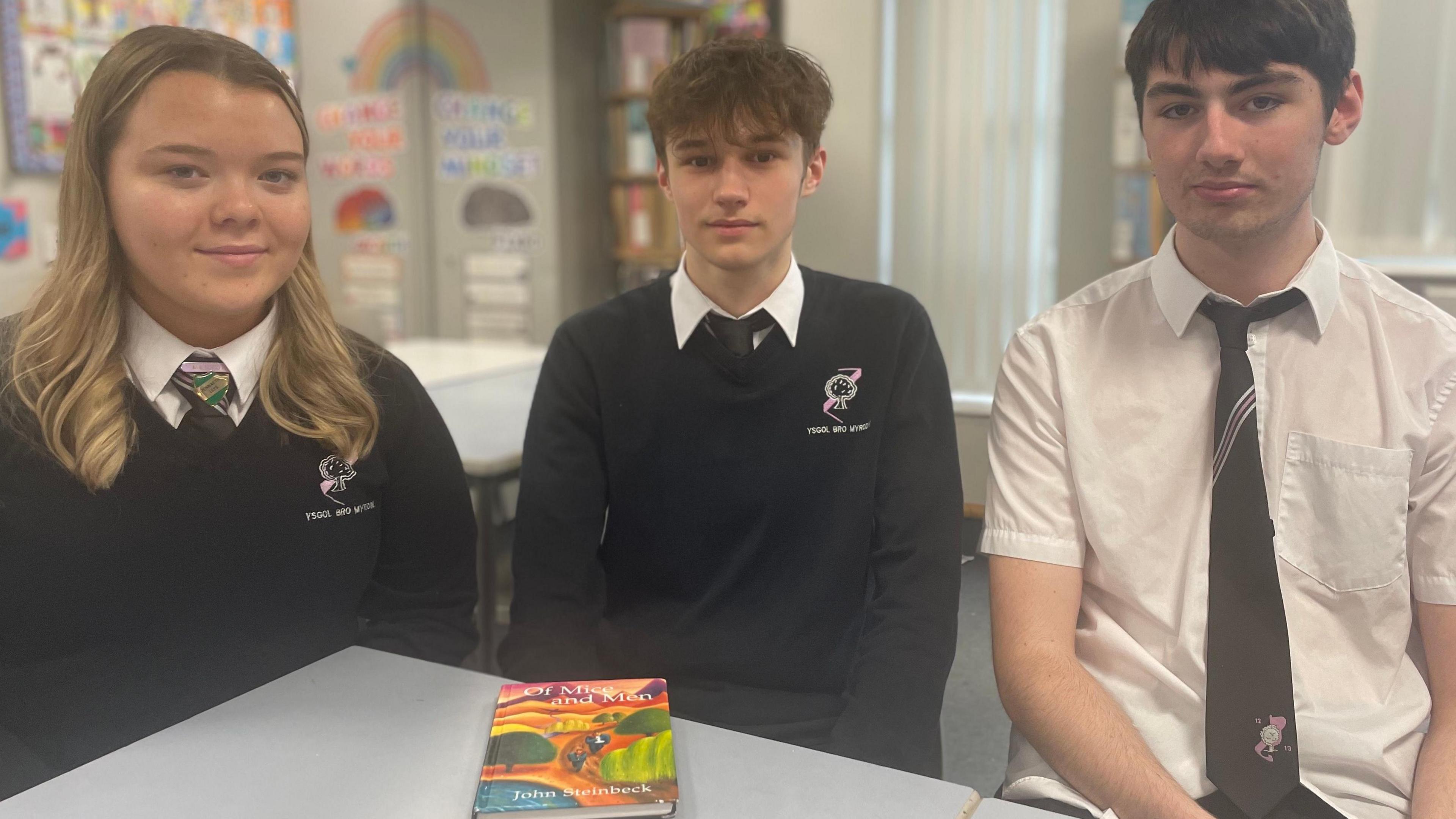 Three pupils - a girl and two boys - in school uniform sitting next to each other in a classroom with a copy of Of Mice and Men on the table in front of them