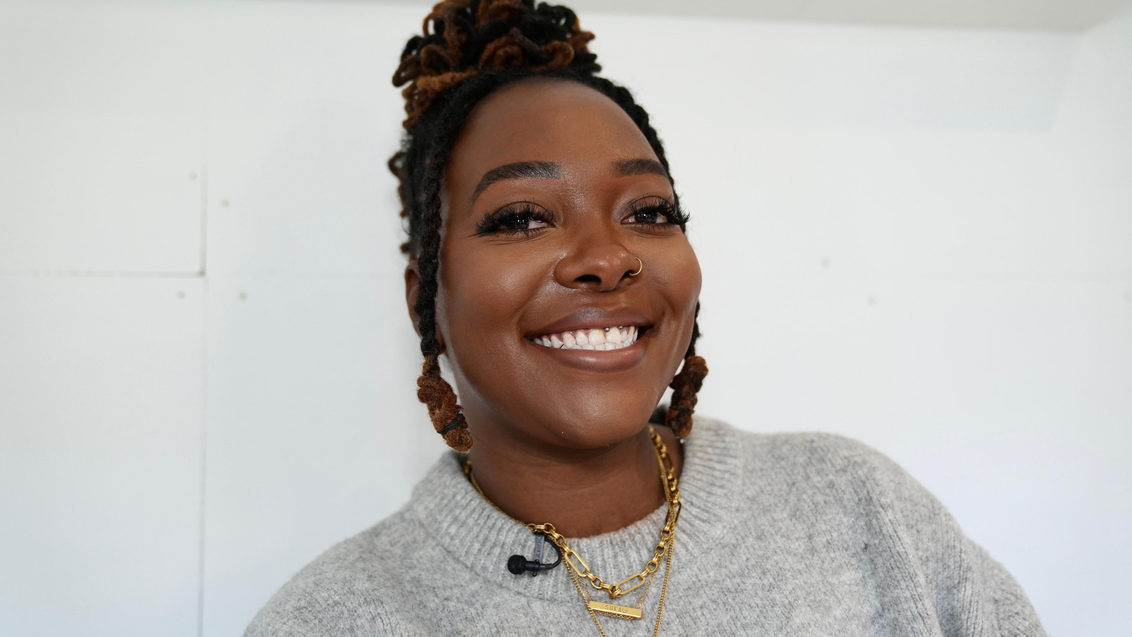Shea Osei smiles at the camera. She has black hair in twists and a ring in her nose. She is wearing a grey sweater and is standing in front of a white wall.