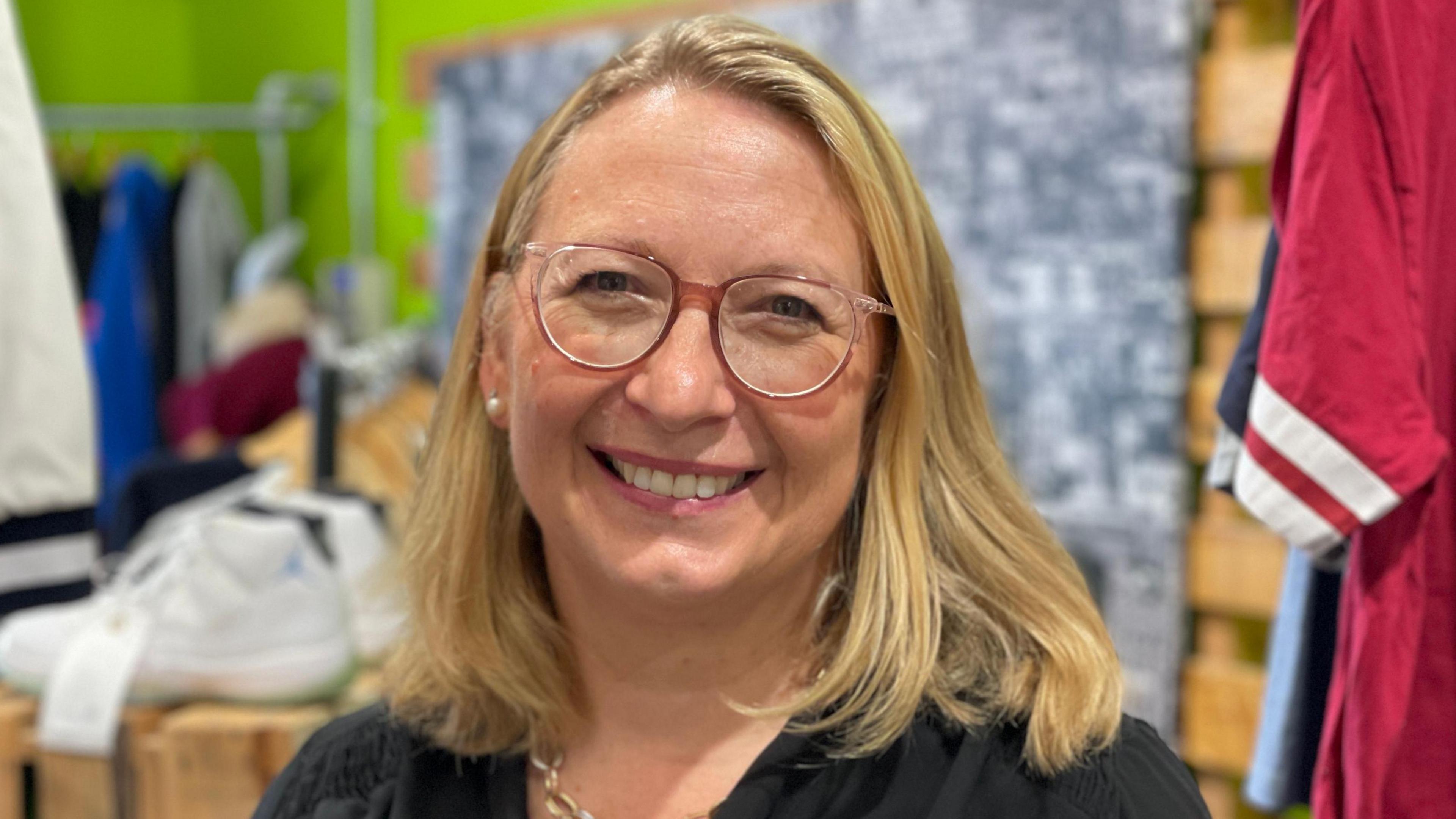 A close-up of Judie Newman from St Elizabeth Hospice. She is wearing glasses and smiling directly into the camera. Tops and a pair of trainers can be seen behind her