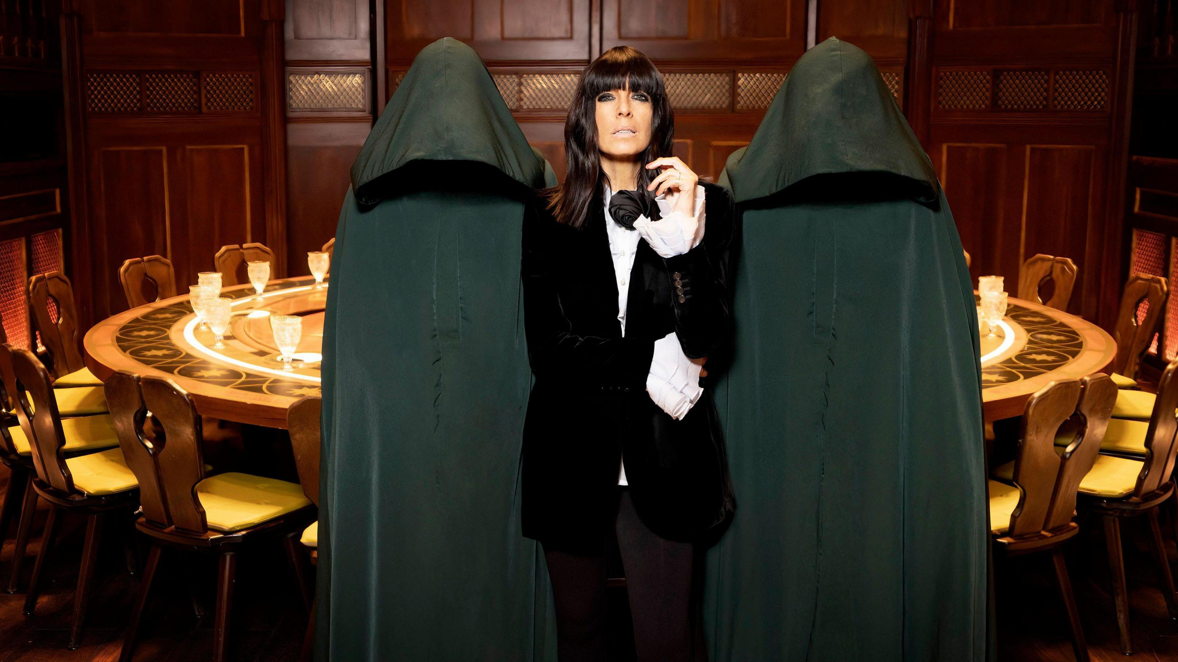 Claudia Winkleman wears a smart black jacket over a white shirt in a promotional photograph for the BBC series Traitors. She stands in front of a table and two hooded figures.