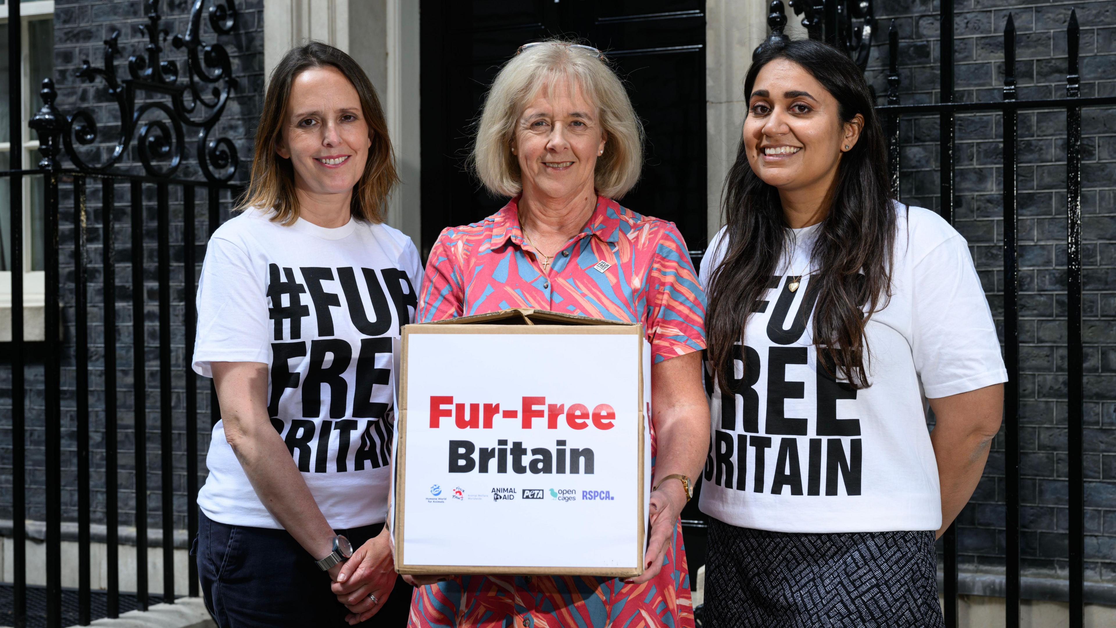 Ruth Jones stands beside two women wearing white shirts that say #FurFreeBritain. Ms Jones is holding a cardboard box with the same words on the outside. She is smiling and is wearing a pink and blue patterned shirt. She has shoulder-length blond hair. The woman to her left has shoulder-length brown hair and is smiling. The woman to Ruth's right has long dark-brown hair and is also smiling. The trio are stood outside Number 10