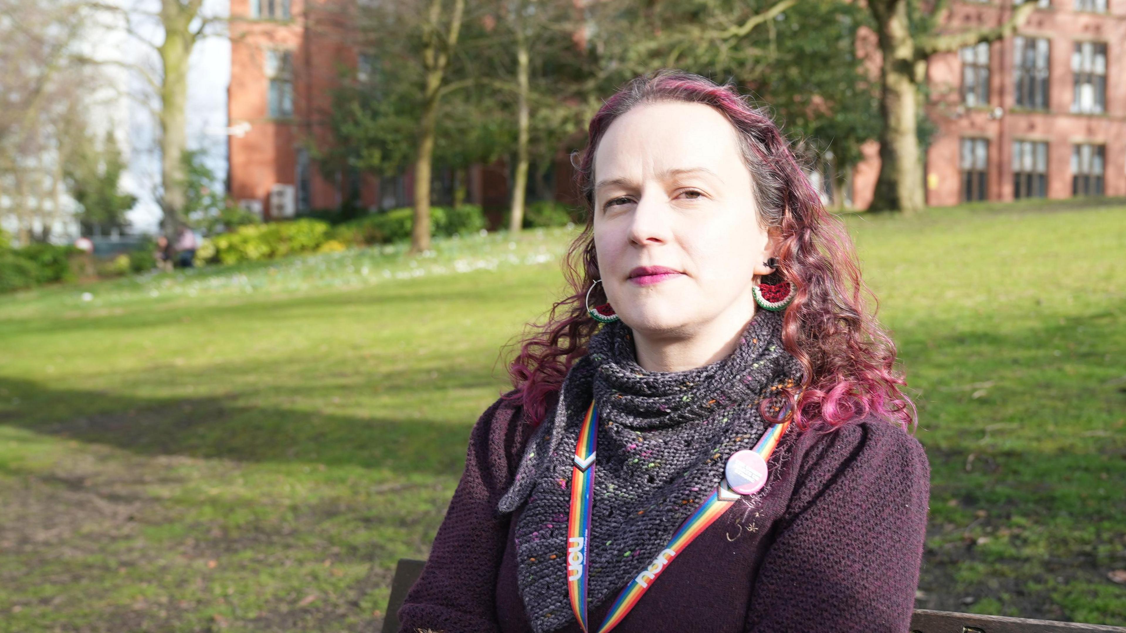 Jenny Hughes looks into the camera dispassionately. She has pink curly hair and is wearing a purple cardigan, with a rainbow lanyard around her neck. She is sat on a park bench with a university building in the background.