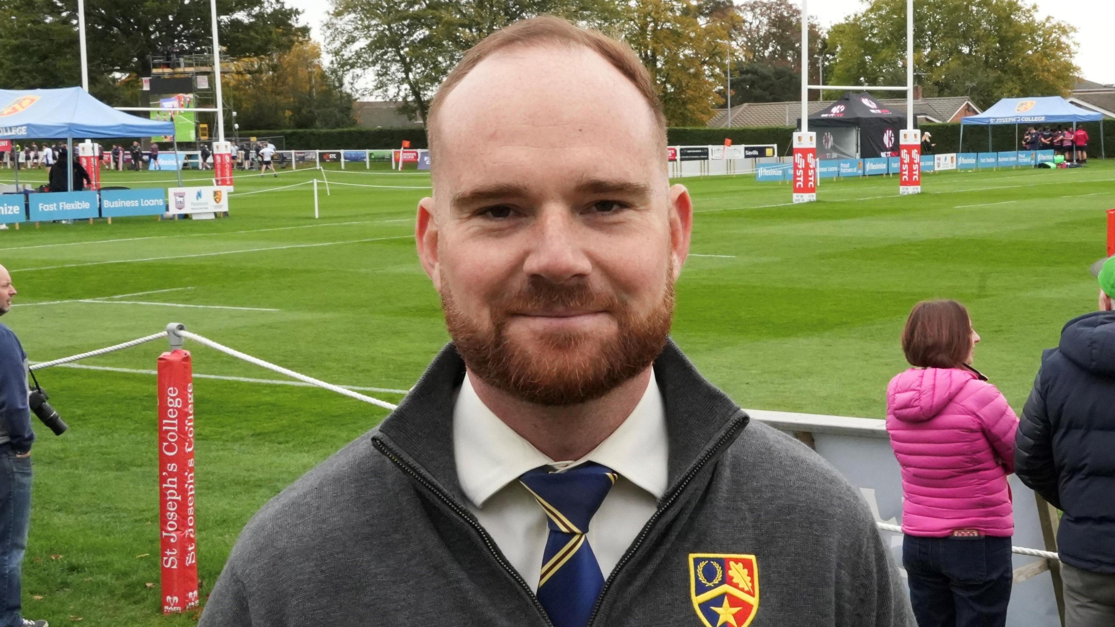 Fred Wenham was standing in the middle of the image looking towards the camera with a light smile. He was wearing a grey zip up jacket and was wearing a white T-shirt and blue tie. He was standing on a green field.