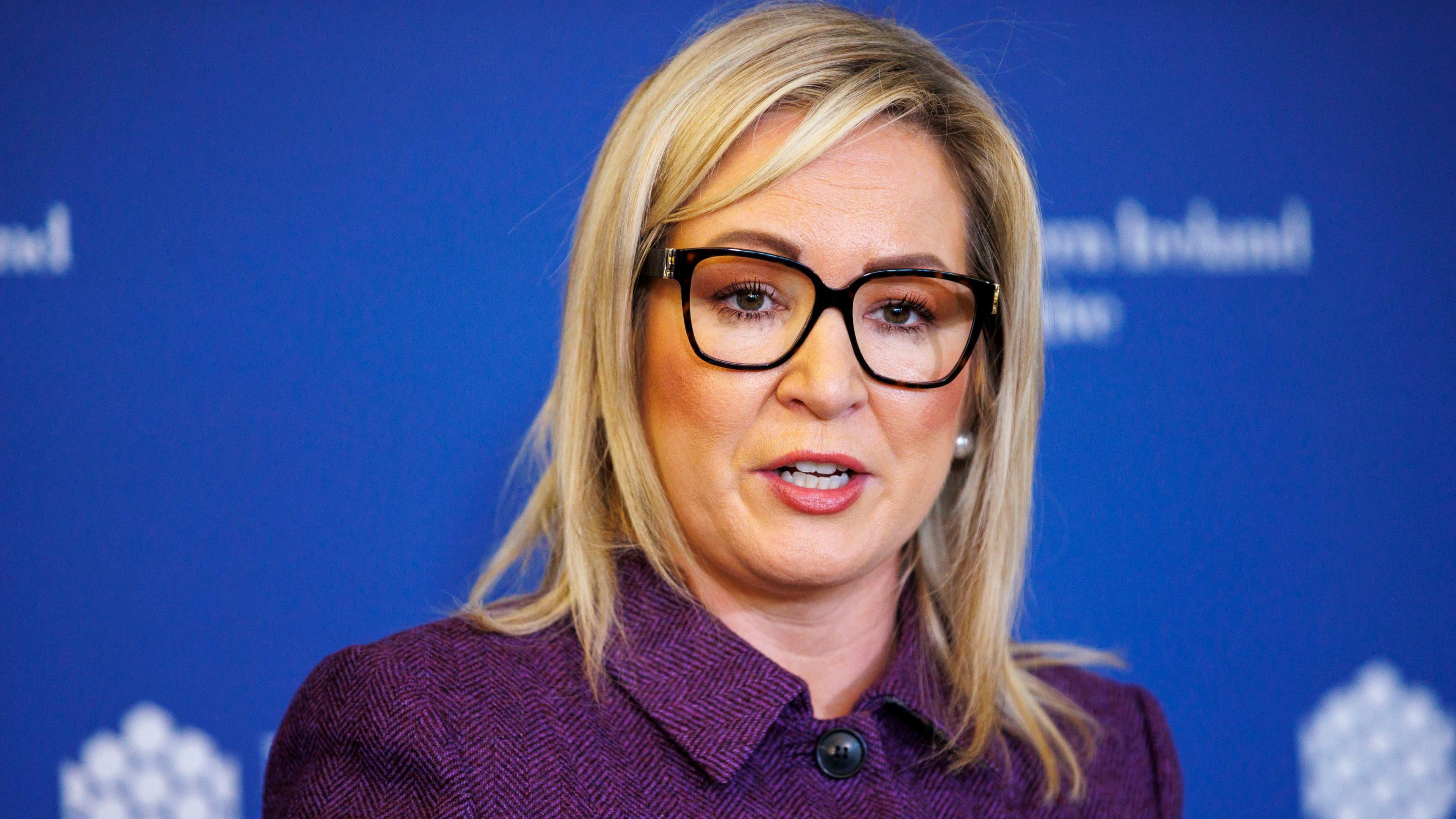 A woman with blonde hair wearing black glasses and a purple coat.