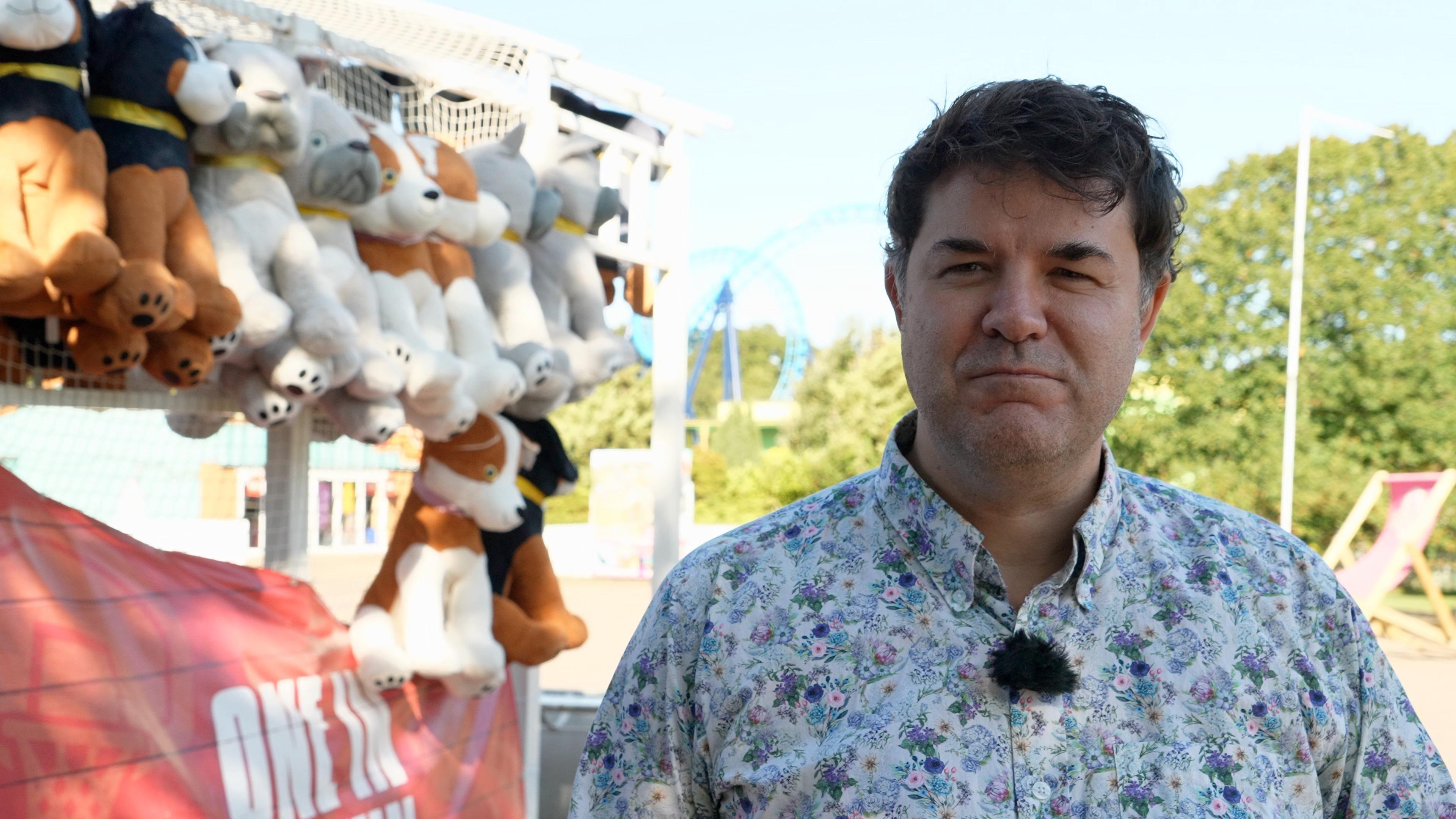 A man wearing a shirt with a floral pattern is stood in the middle of a theme park near some stuffed toys