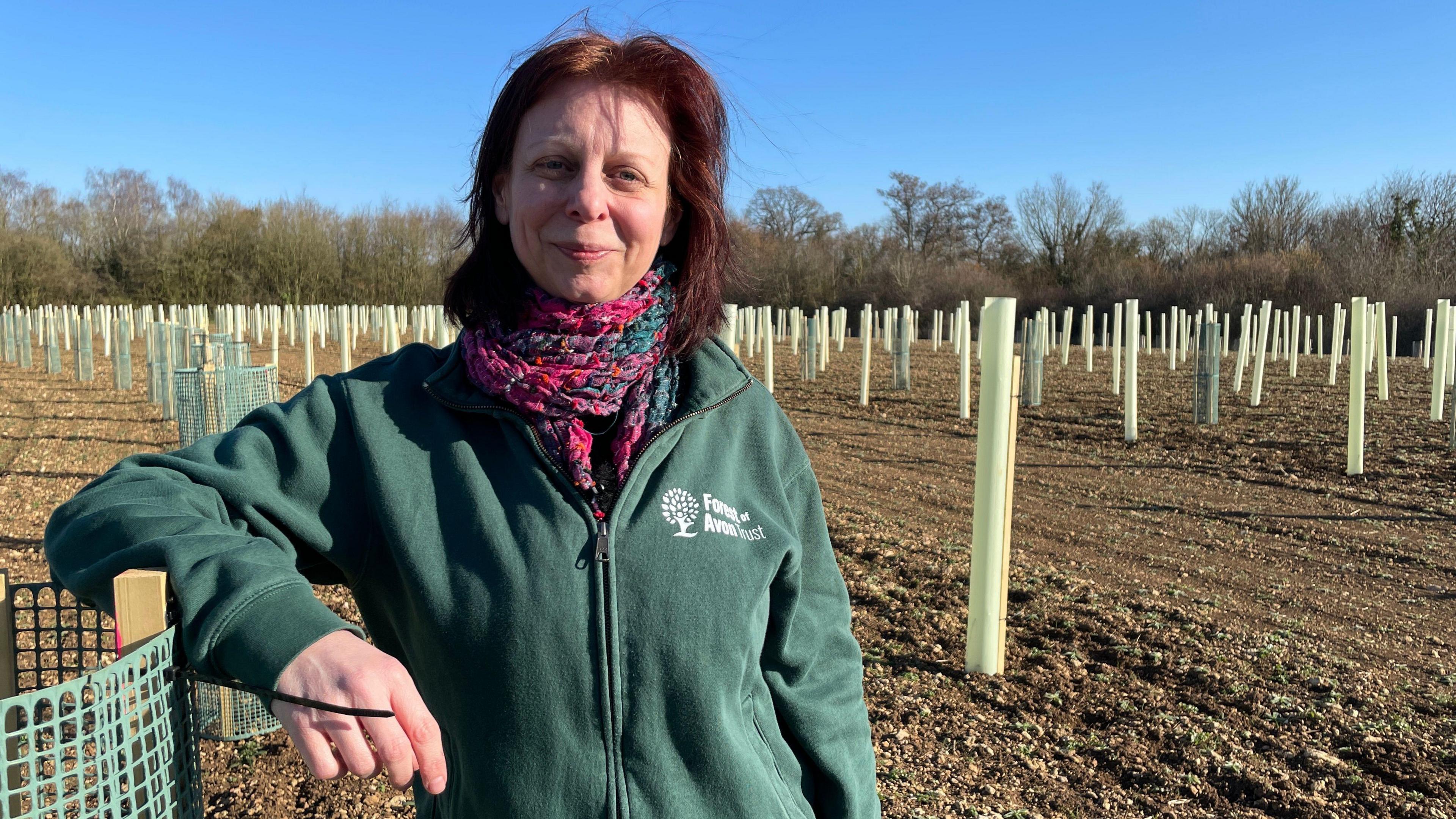 Alex Stone, chief executive of the Forest of Avon Trust, stands in a field filled with newly-planted trees. She is leaning on a plastic guard used to protect the saplings.