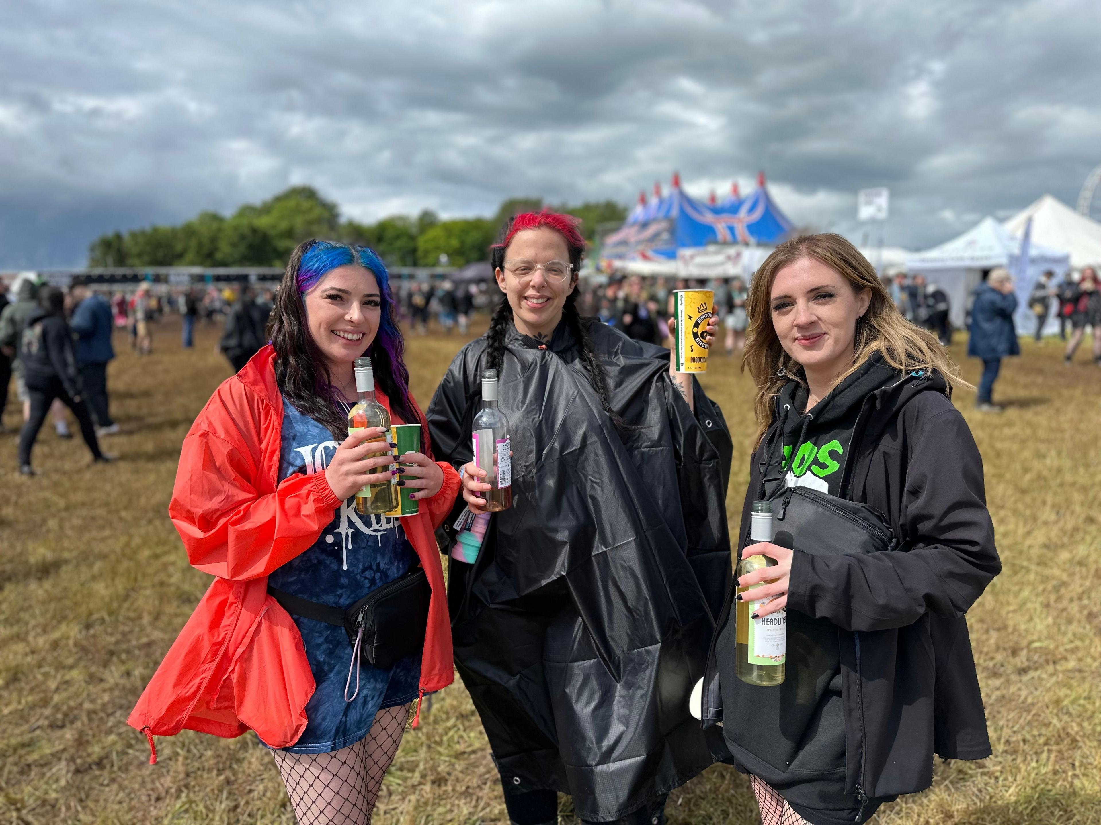 Three girls stood in a field in front of a tent, holding drinks and wearing ponchos. The sun is shining but it is cloudy.