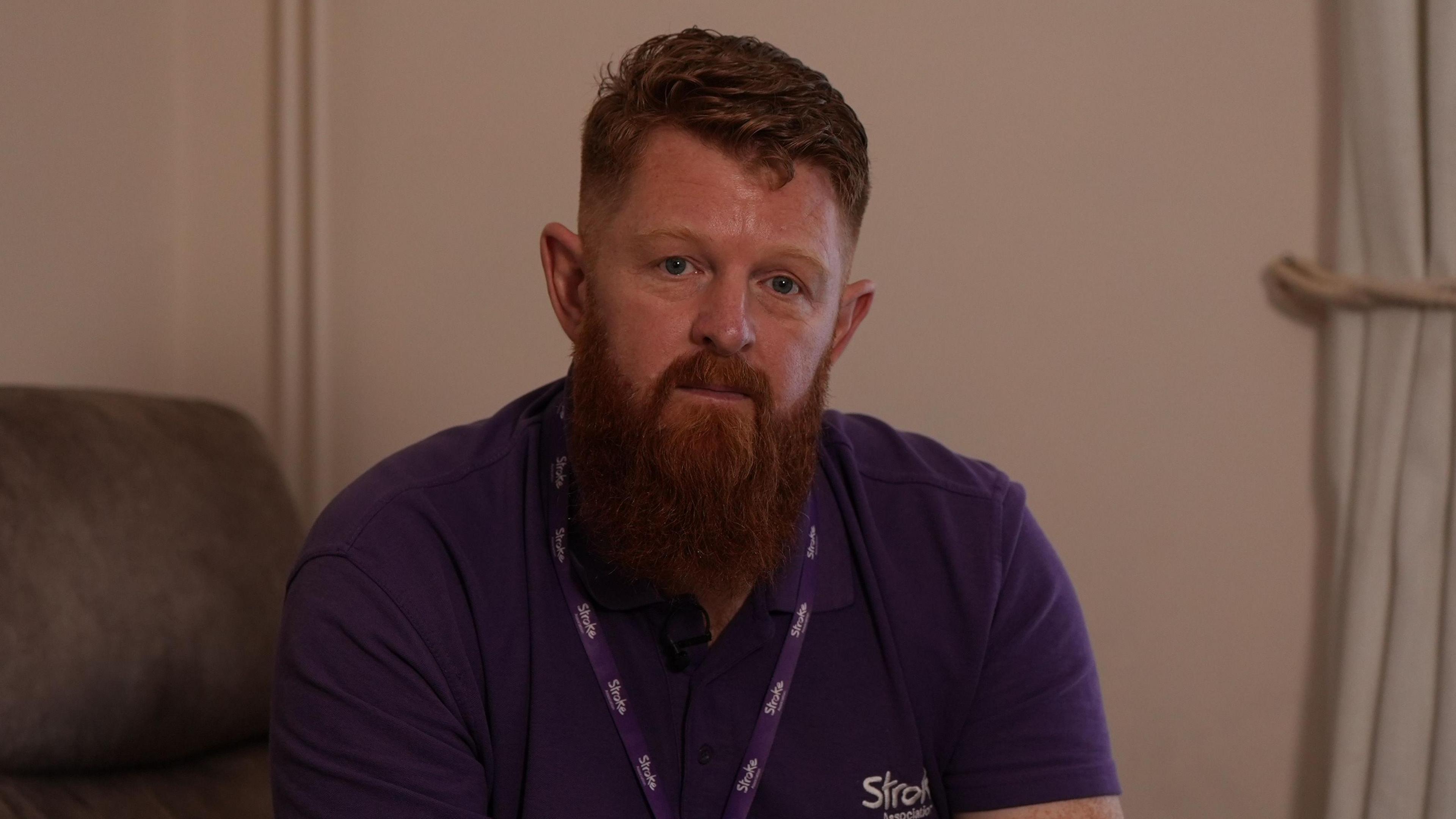 Dave Jones has a red beard. He is perched on the edge of a sofa and wears a purple Stroke Association t-shirt and lanyard. 