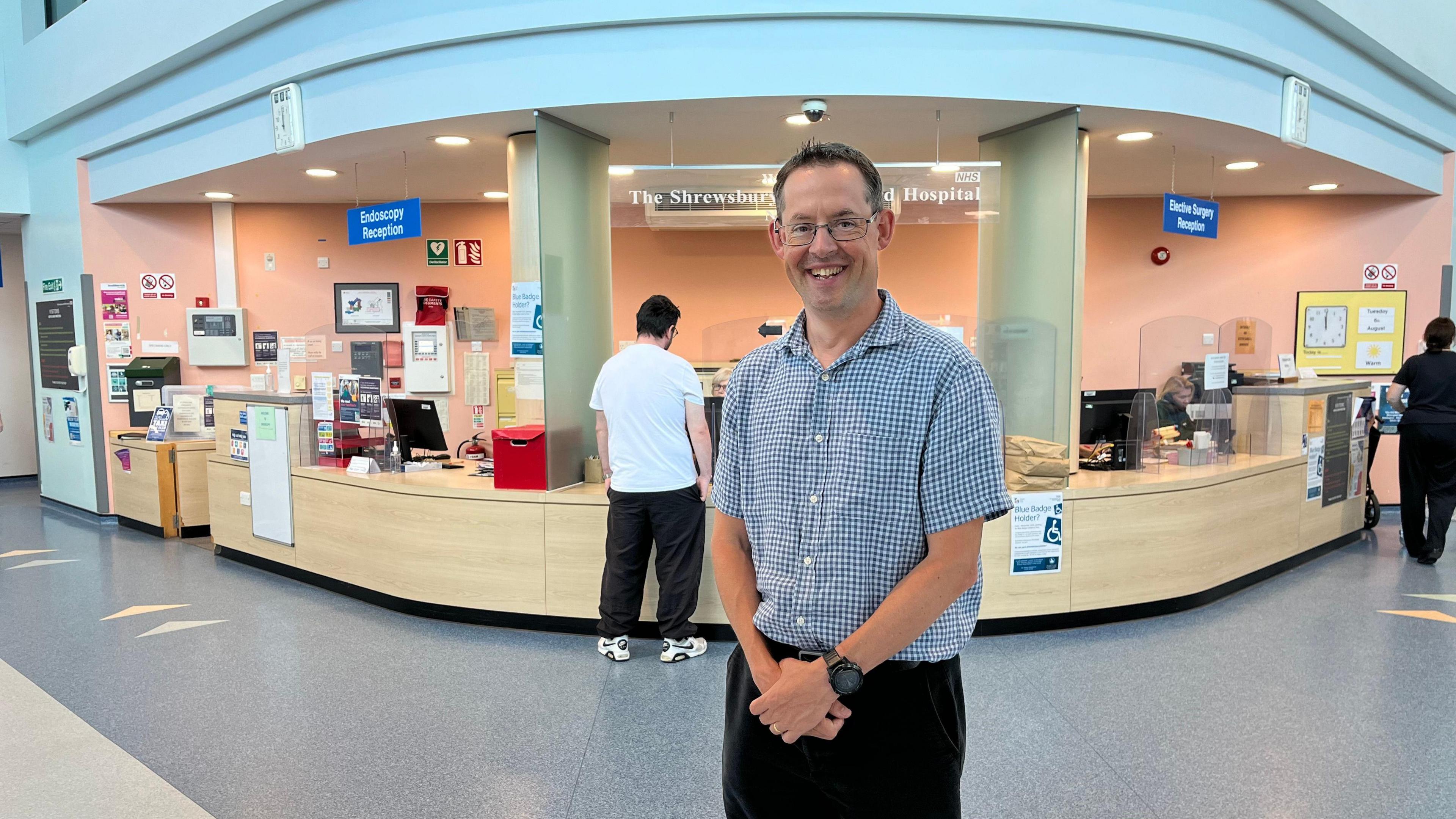 Ed Rysdale is dressed in a blue and white checked shirt. He has glasses and dark hair, and is smiling. He is standing in a hospital entrance area.