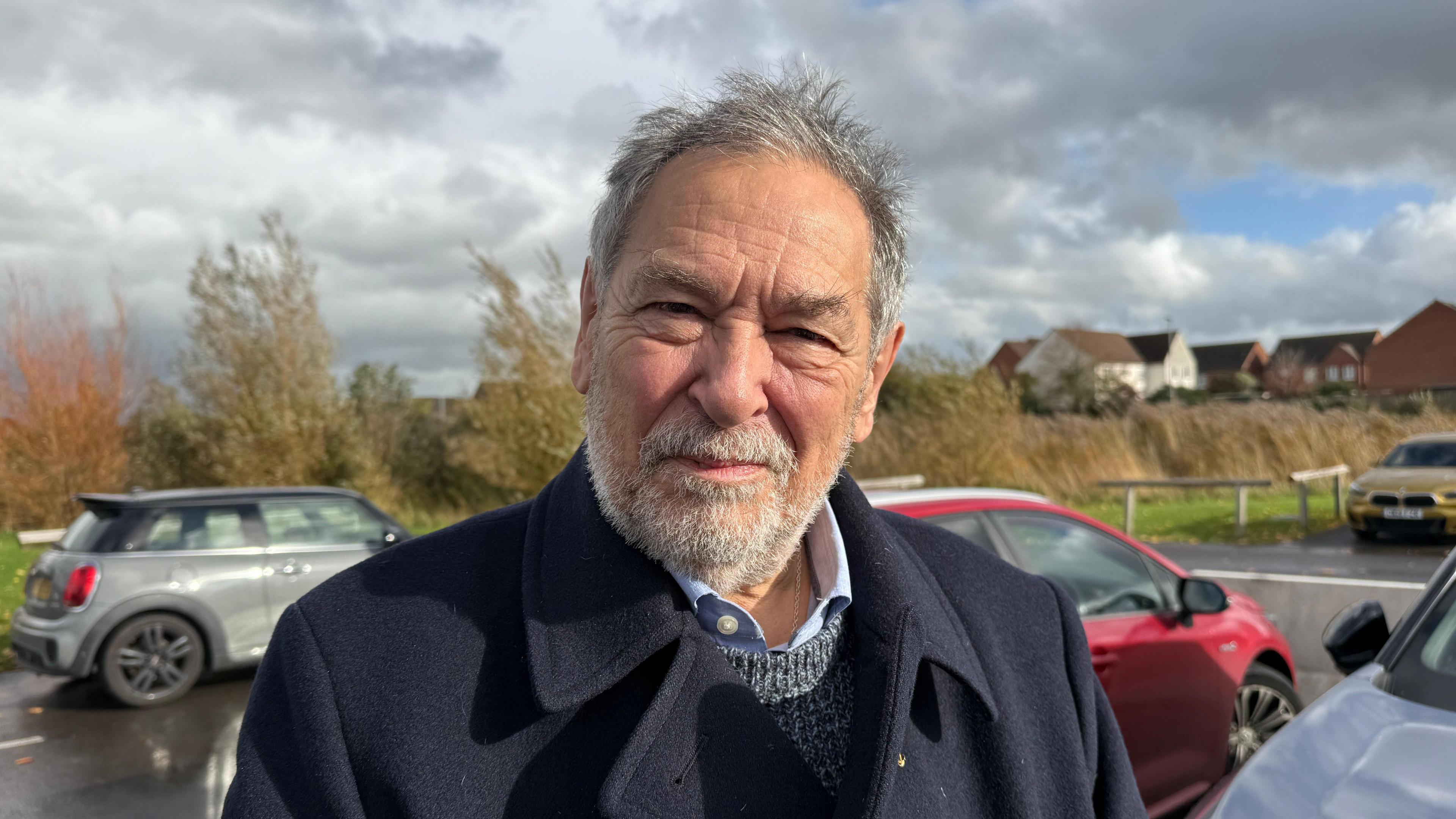 Mike Caswell is wearing a navy coat.  He has grey hair and a grey beard.  He is standing in a car park and there are houses in the background.
