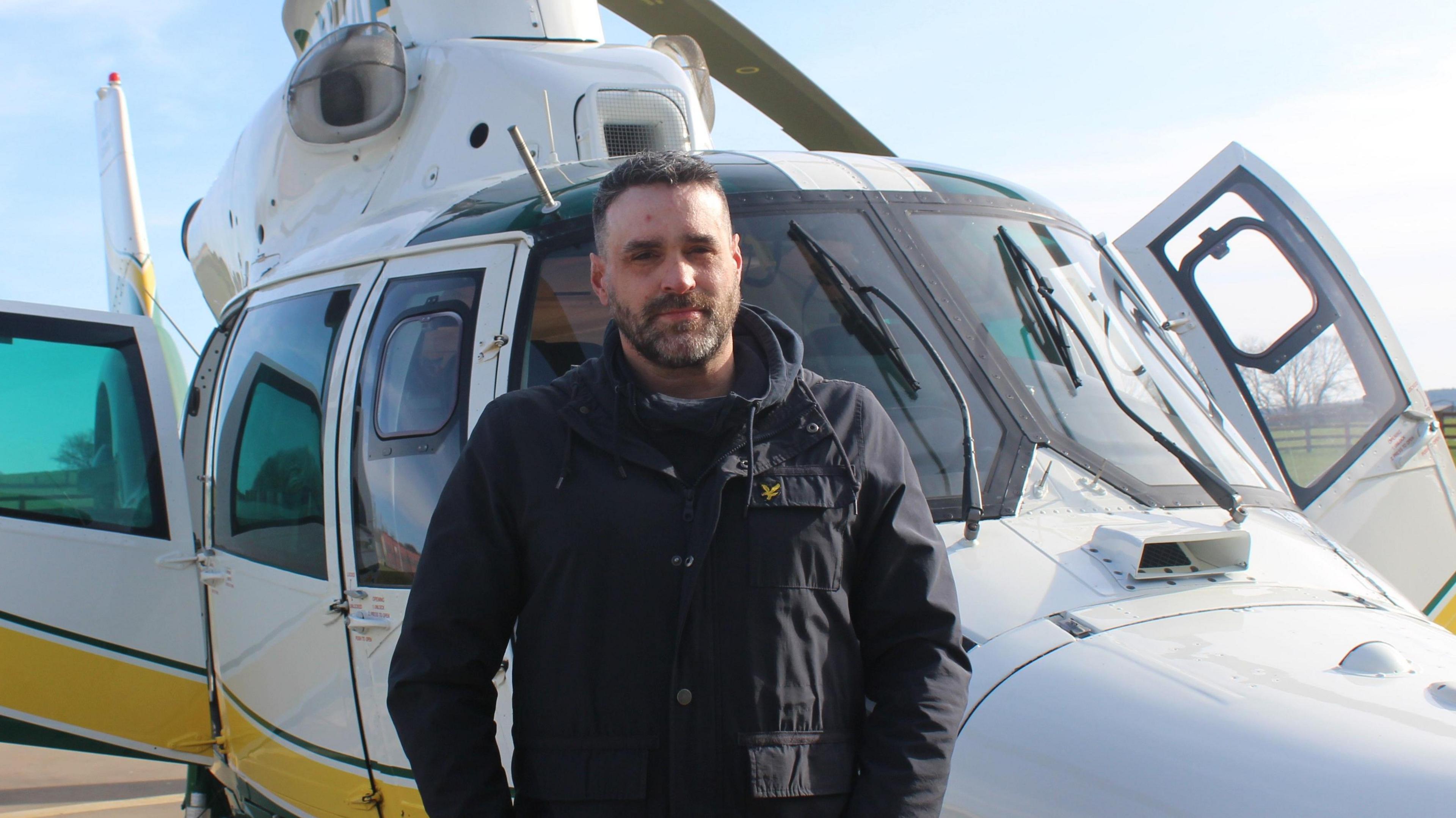 Mr Taylor standing in front of the GNAAS air ambulance. He is wearing a black coat. He has short brown hair and a short beard. His face has recovered from the burns he suffered in the blast.