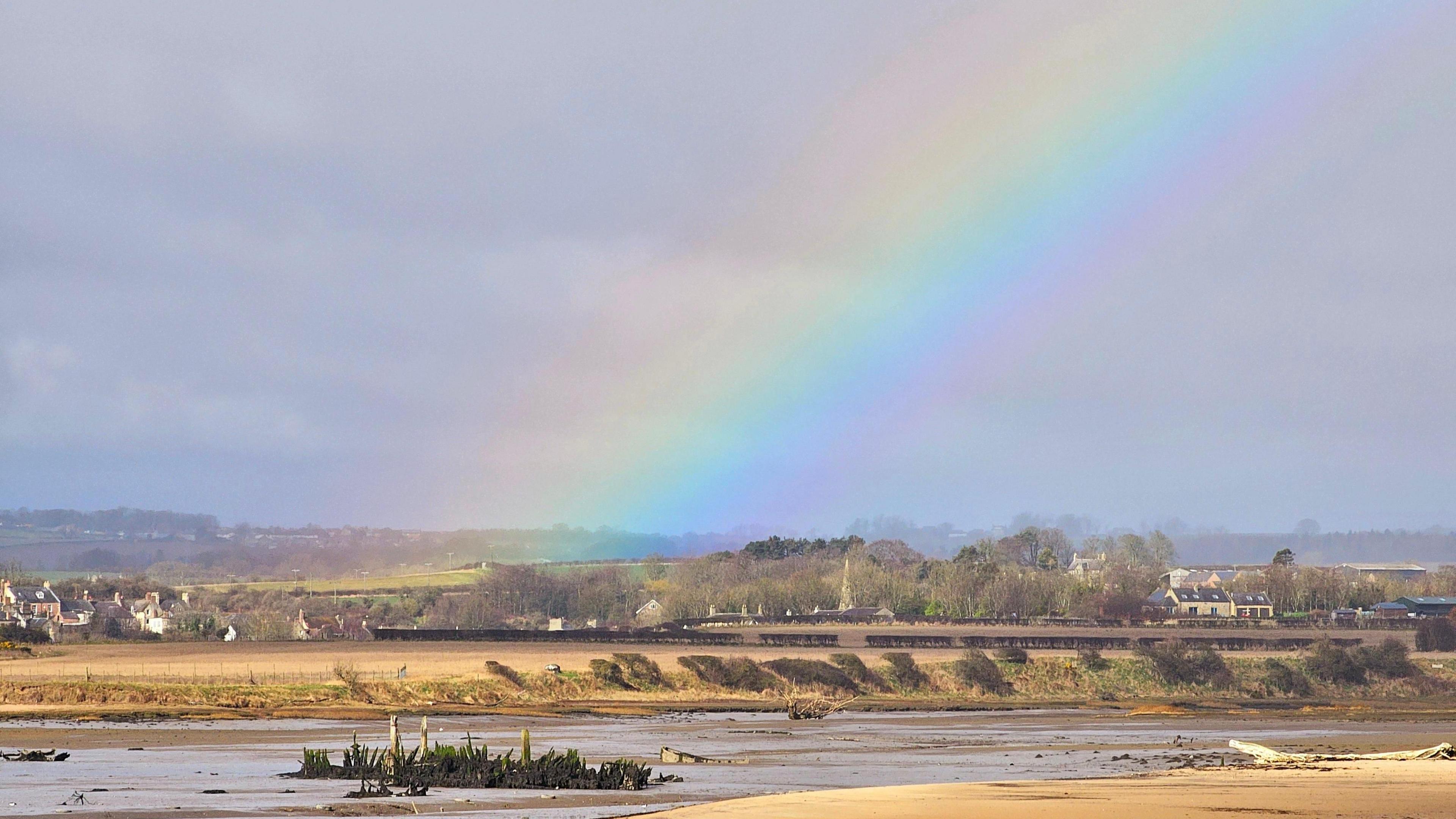 A rainbow over a sandy beach. Rural fields and countryside houses can bee seen in the background.