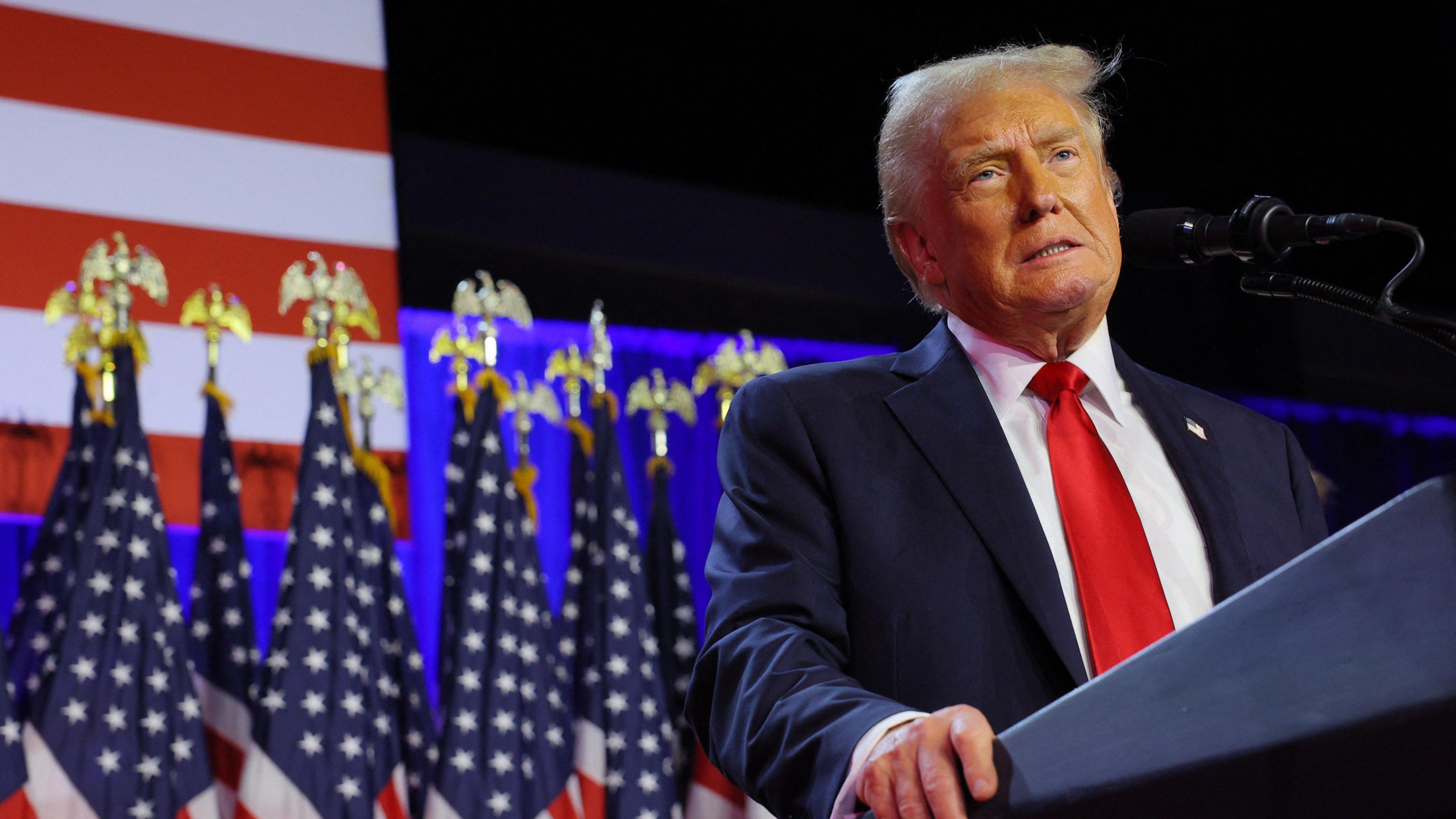 Donald Trump at a lecturn speaking into a microphone in front of multiple American flags. Trump is wearing a navy suit and red tie