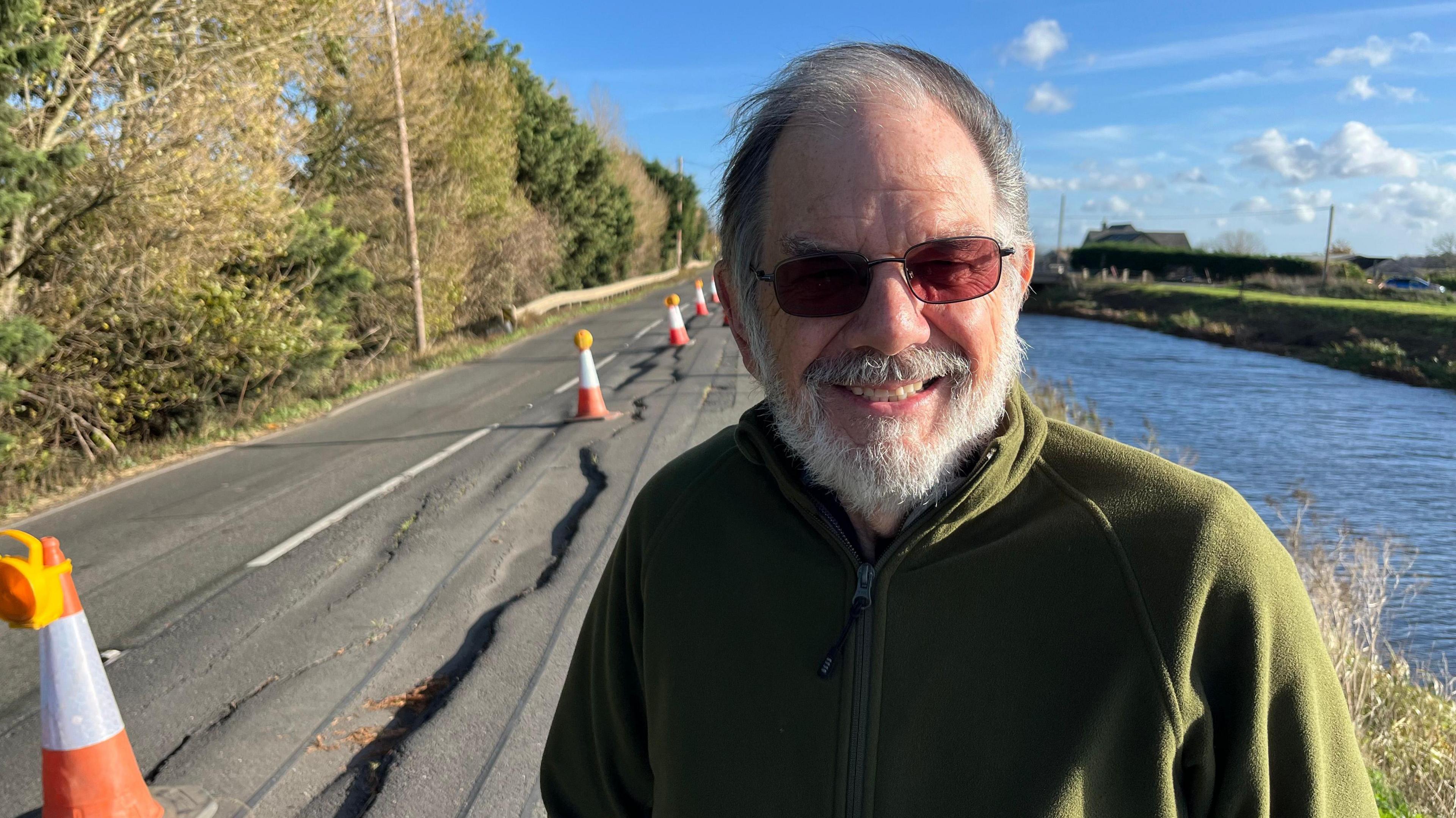 Alan Whitney, 76, has a grey beard and moustache. His black and grey hair is being blown across his head by the wind. He has glasses on with a tint on the lenses and he is wearing a bottle green fleece. Behind is a badly cracked road with orange and white cones near the middle white lines. On the right is a waterway.