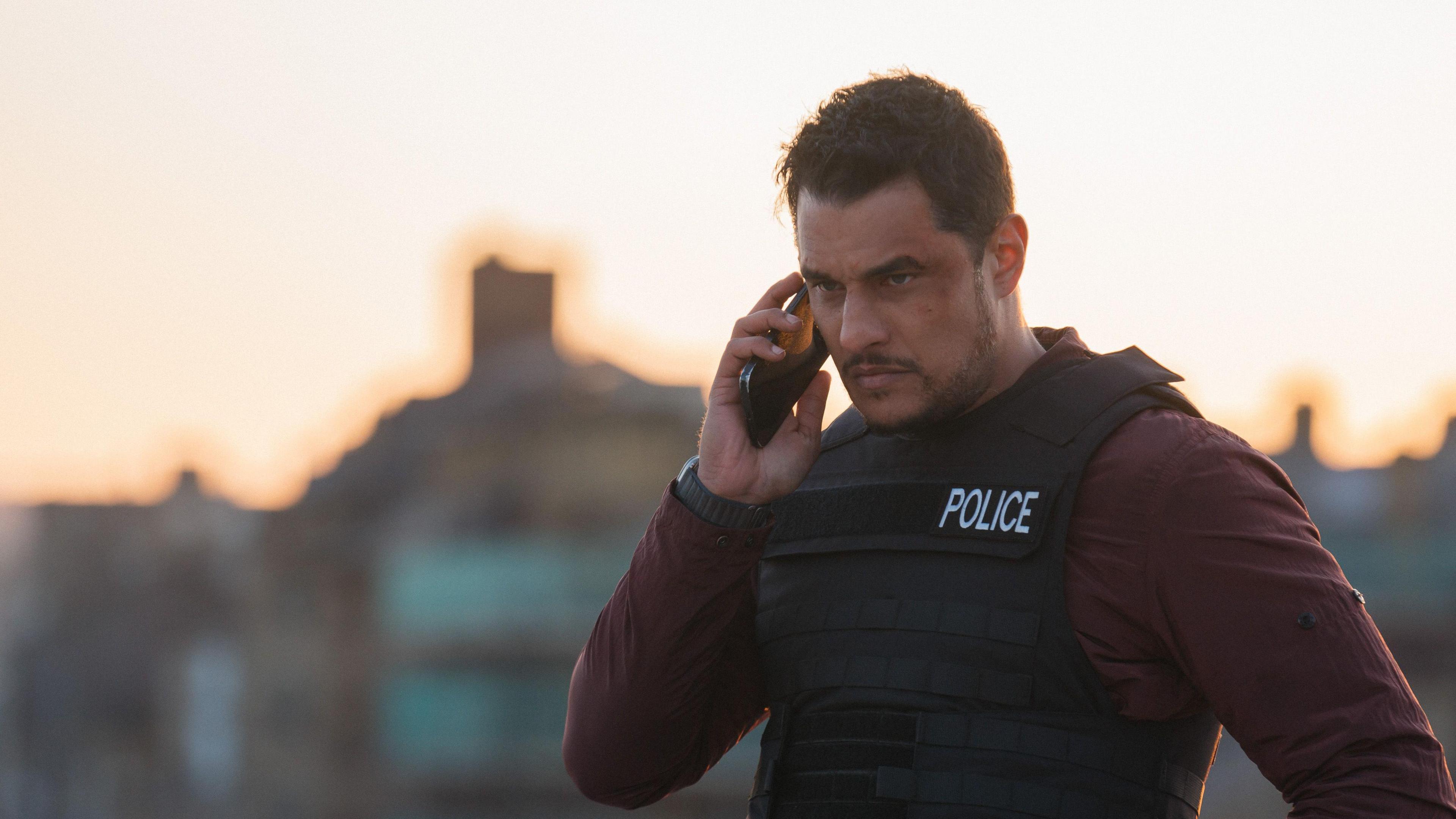 Actor Staz Nair as Detective Harry Virdee, wearing a police-labelled bulletproof vest and taking a phone call.