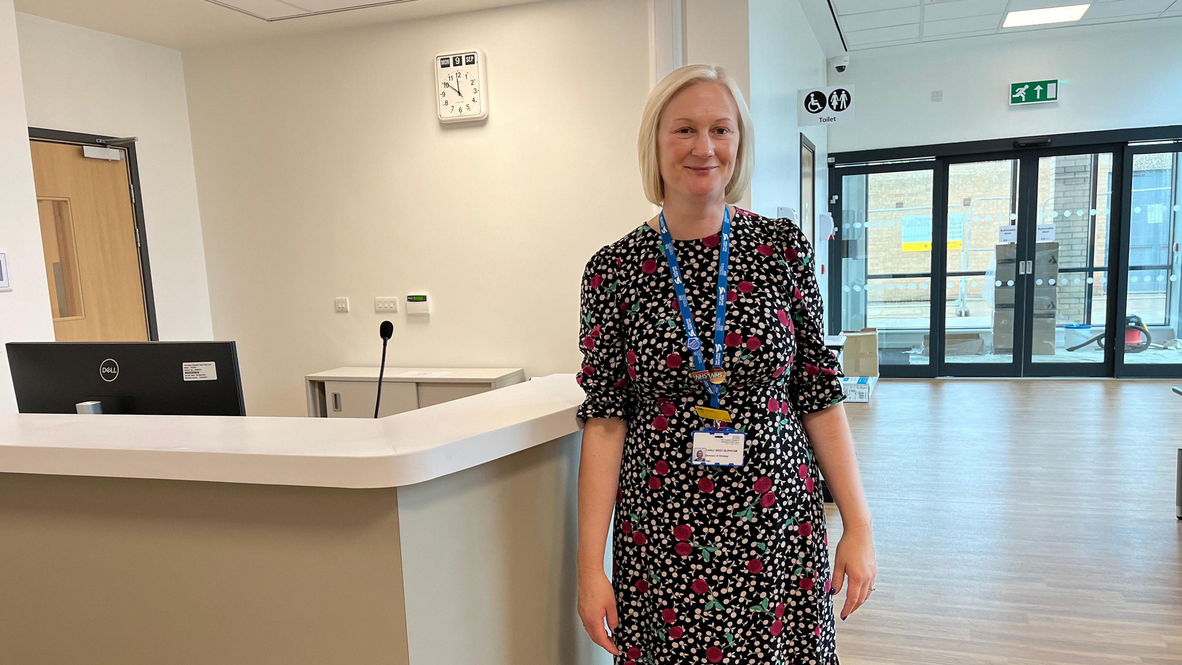 Carly West-Burnham the hospital's director of strategy stands in front of the new reception desk inside the Community Diagnostic Centre. She is  wearing a black dress decorated with red cherries and white blossom. She has a blonde bob and is wearing and NHS ID card around her neck. 