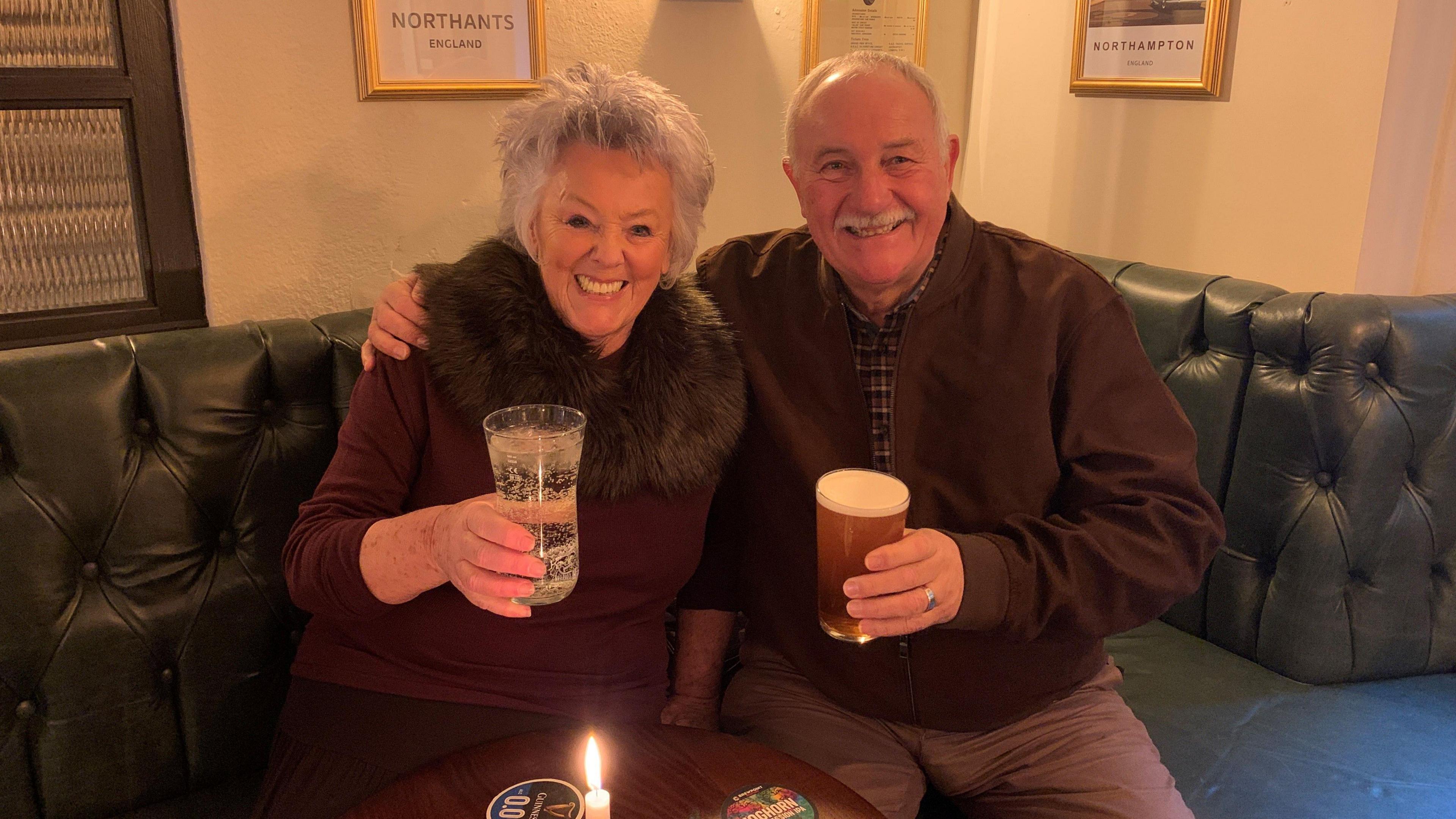 A woman and a man in their 70s beside each other in a pub, with their glasses raised. The woman has silver hair, with a fur garment around her shoulders and a burgundy sweater. The man has grey hair and a moustache and his arm around the woman. 