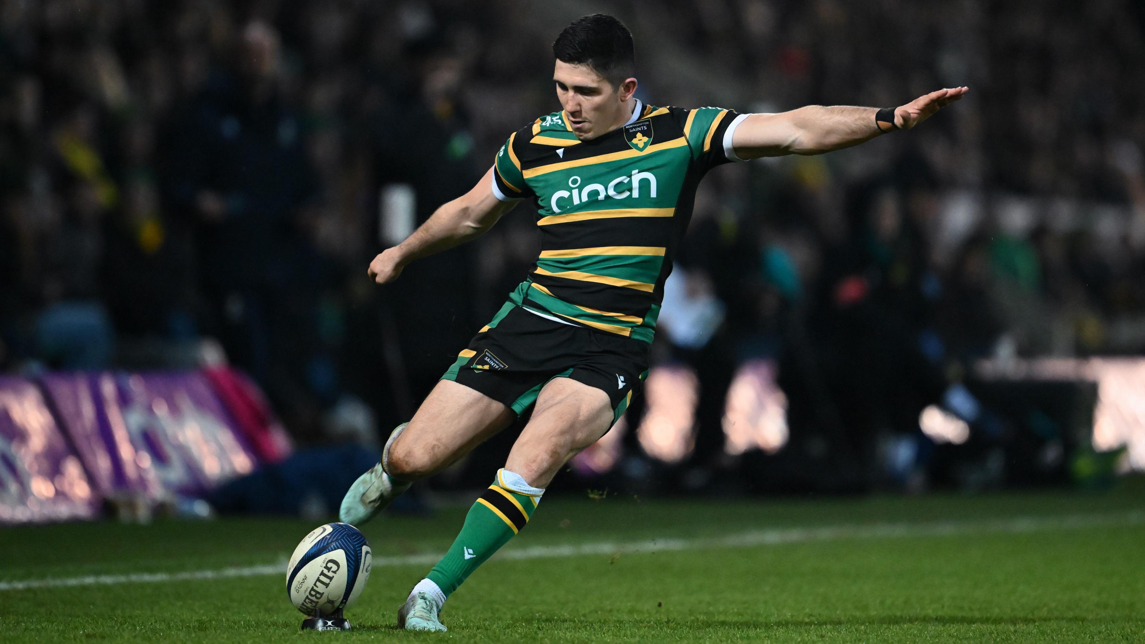 Anthony Belleau holds one arm out to his left and swings his right leg back as he takes a kick at goal wearing Northampton Saints home kit