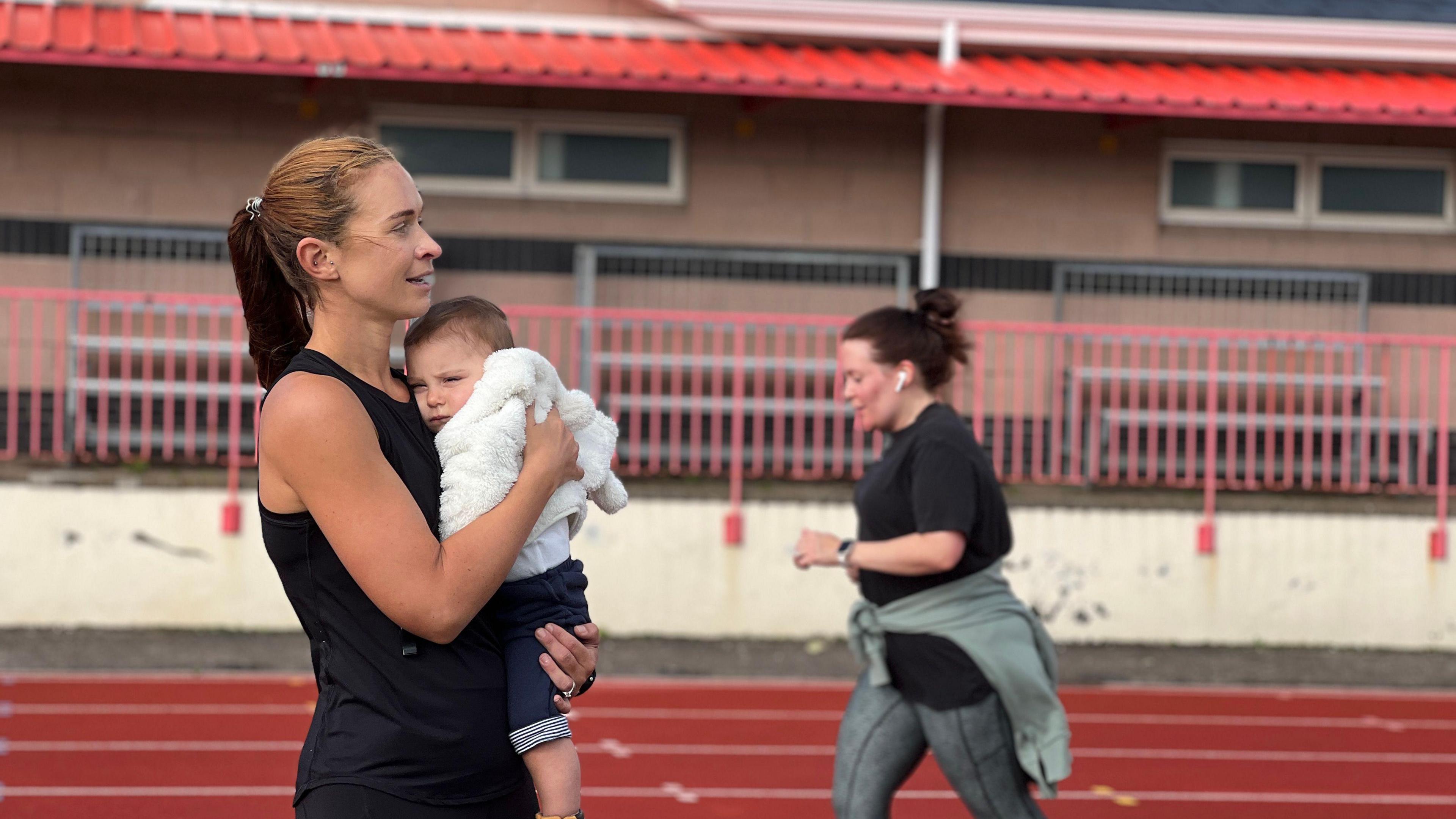 The coach holds a baby as she looks to right. Behind her is a running track and a mum is running behind her to her right. The coach is in black exercise kit and her hair is in a ponytail. The baby is wearing a white coat and blue shorts. The mum running has her hair in a bun and is wearing a black exercise top with grey joggers and has a grey coat tied around her waist.