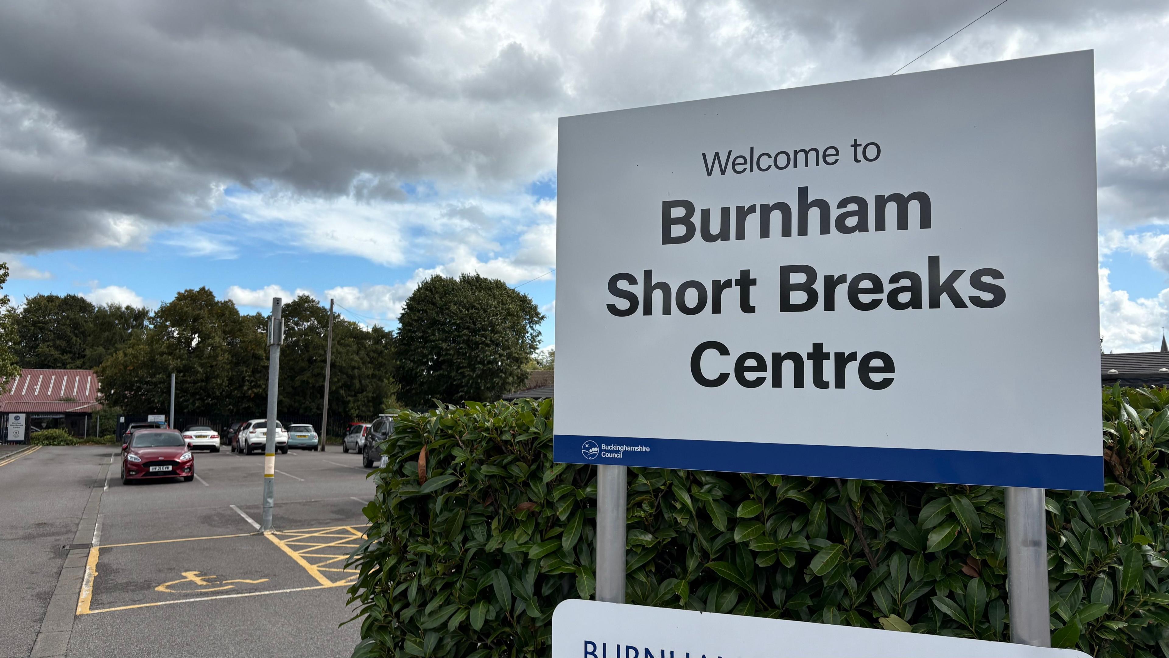 A large sign saying "Welcome to Burnham Short Breaks Centre" in black lettering on a white background. There is a hedge behind the sign, and a car park with yellow lines painted on the ground behind. A single-storey building with a pitched roof is visible on the left in the background.